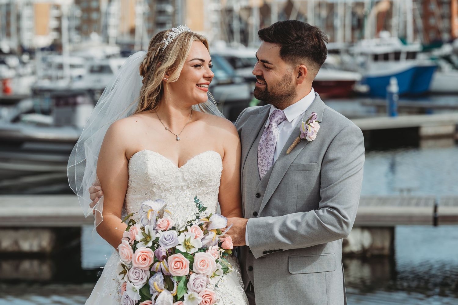 A happy couple shares a joyful moment on their wedding day by a marina with boats in the background.