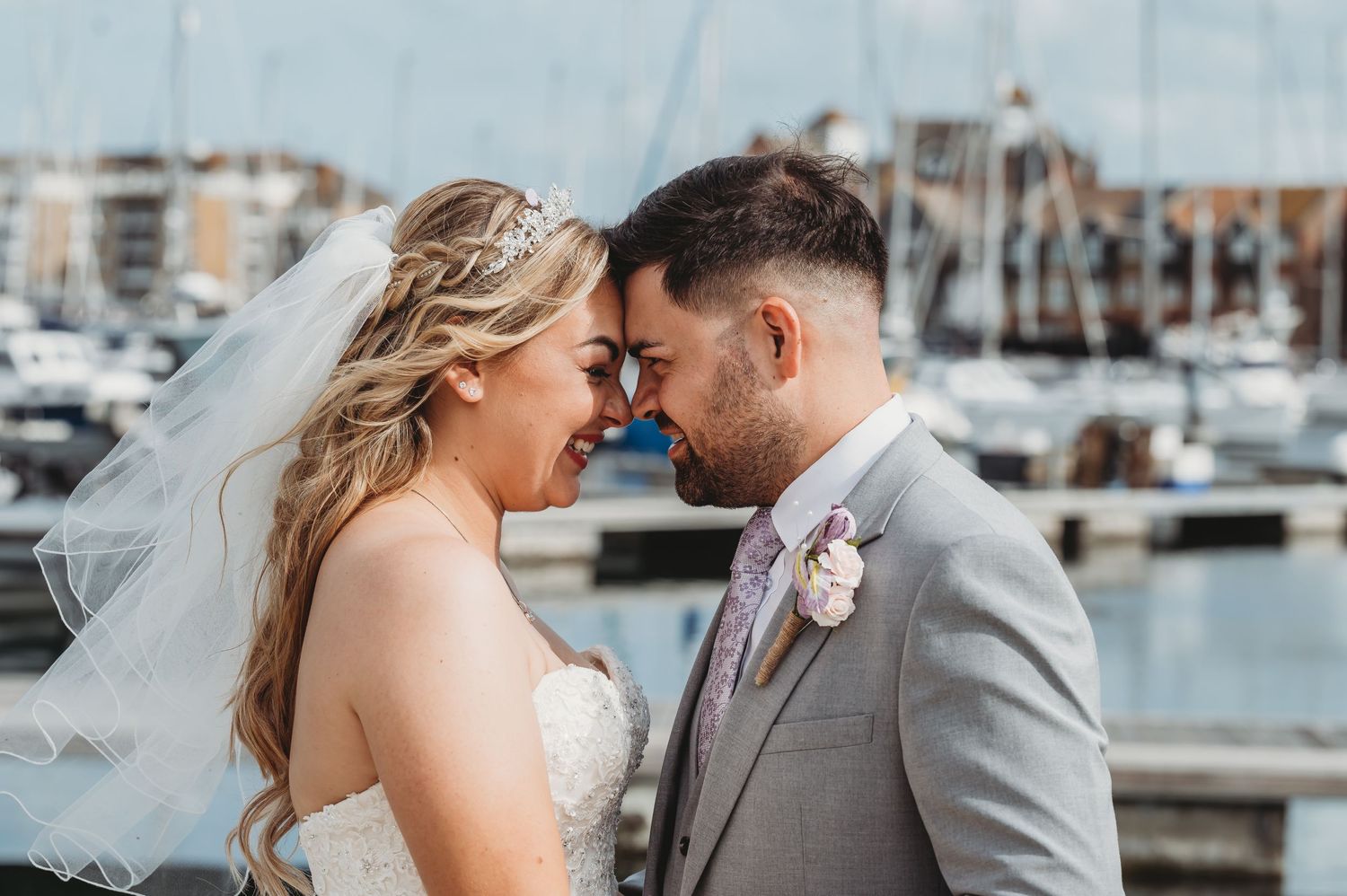 A romantic wedding couple shares an intimate moment at a marina with boats and docks in the background.