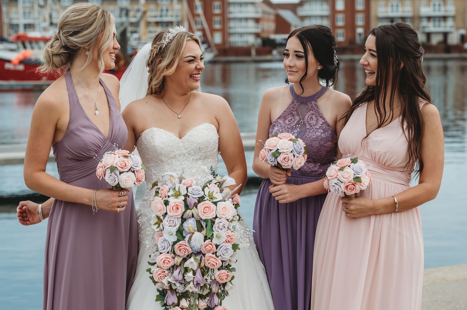 Bride and bridesmaids hold bouquets while laughing together at a waterfront wedding venue.
