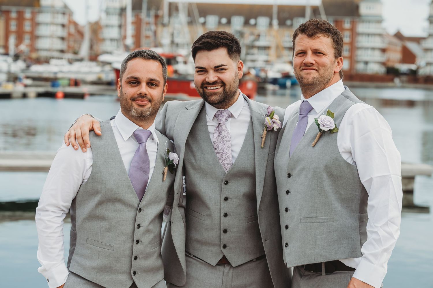 Three groomsmen wearing coordinated gray waistcoats pose together by a waterfront marina.