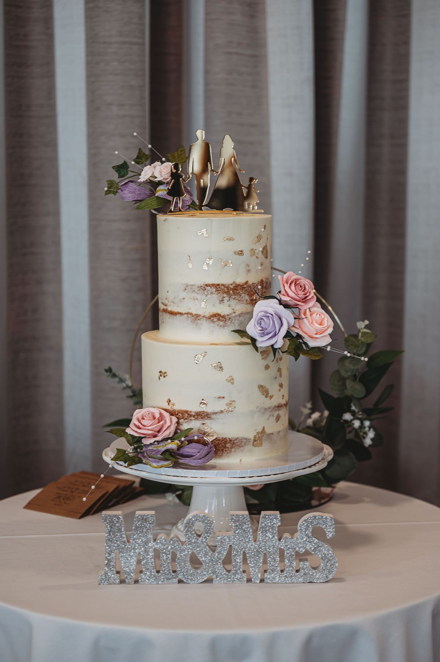 Elegant three-tier semi-naked wedding cake with pink and purple roses on white stand with glittery 'Mr & Mrs' sign.