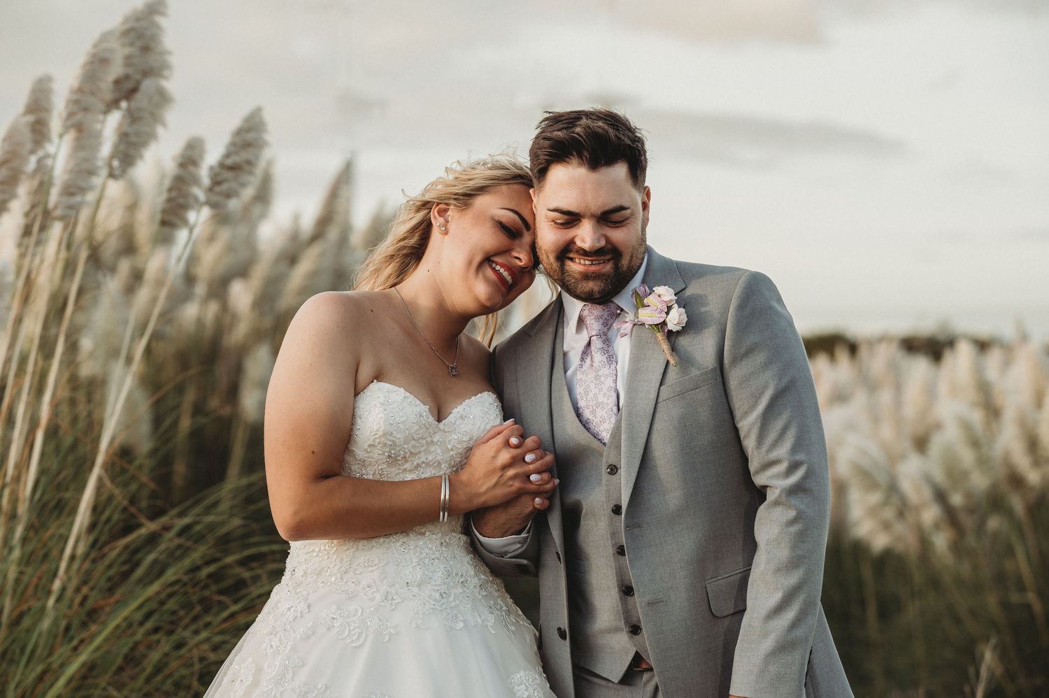 A joyful couple in wedding attire share a candid moment together among tall pampas grass at sunset.