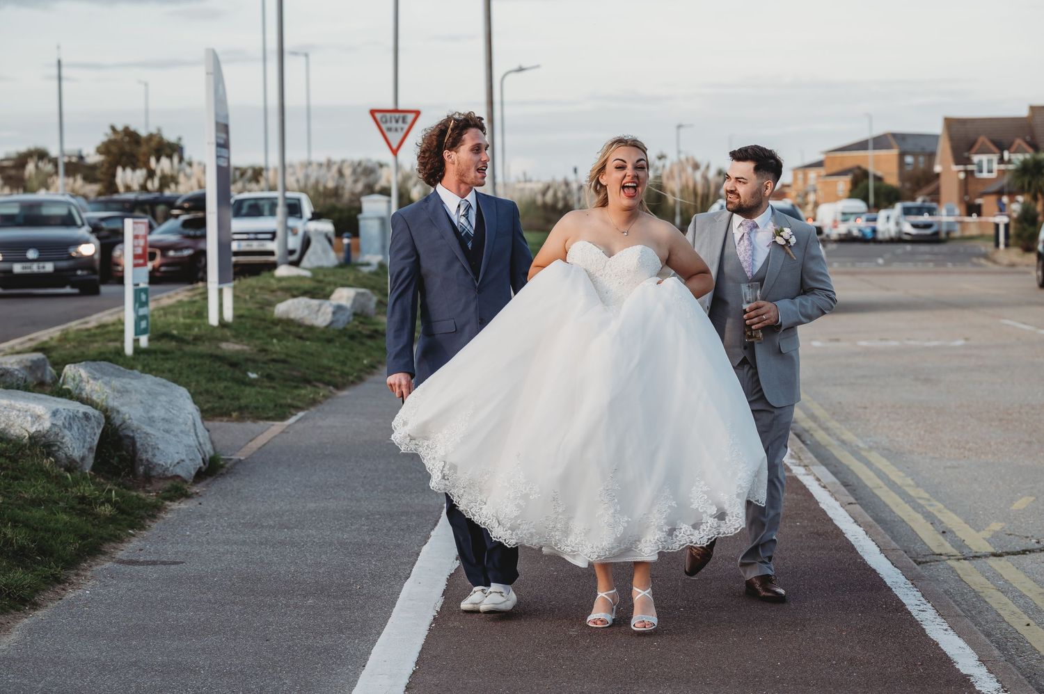 Three people in formal wedding attire walk and laugh together on a sidewalk in a suburban setting.