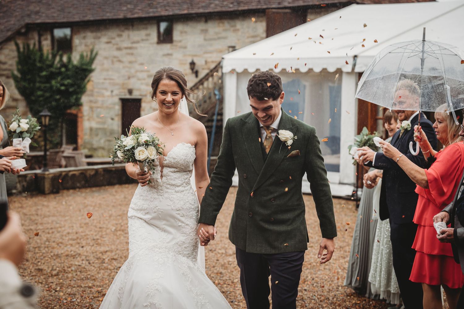 A joyful wedding celebration outside a rustic stone building with guests throwing confetti.