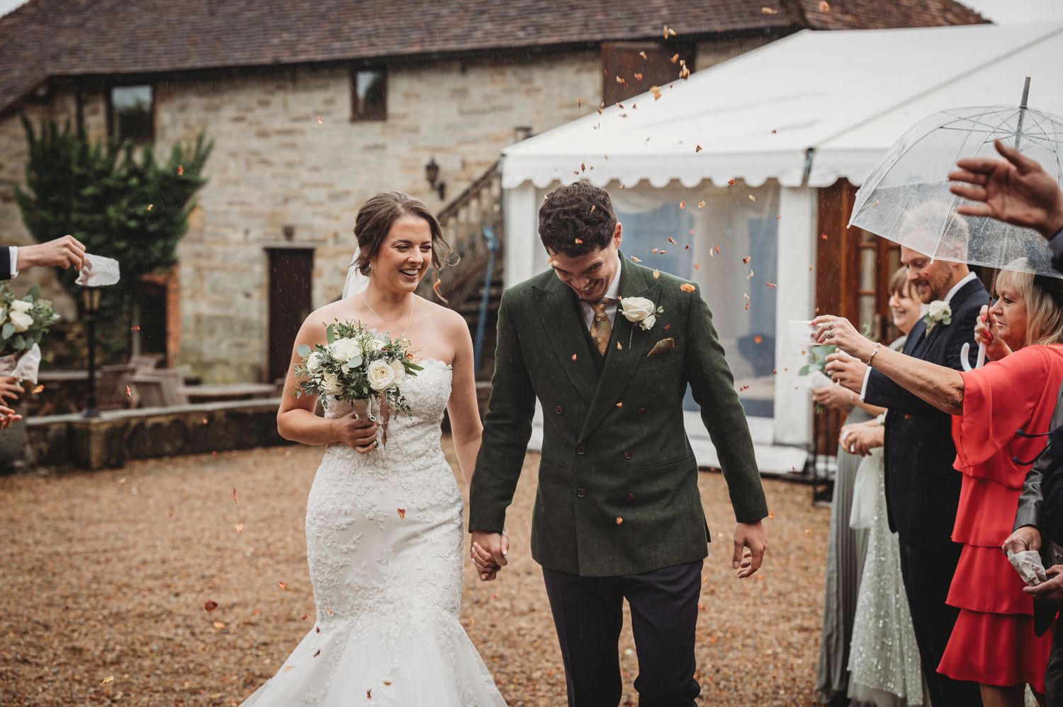 Newlyweds walk together on rustic grounds while wedding guests celebrate with confetti.