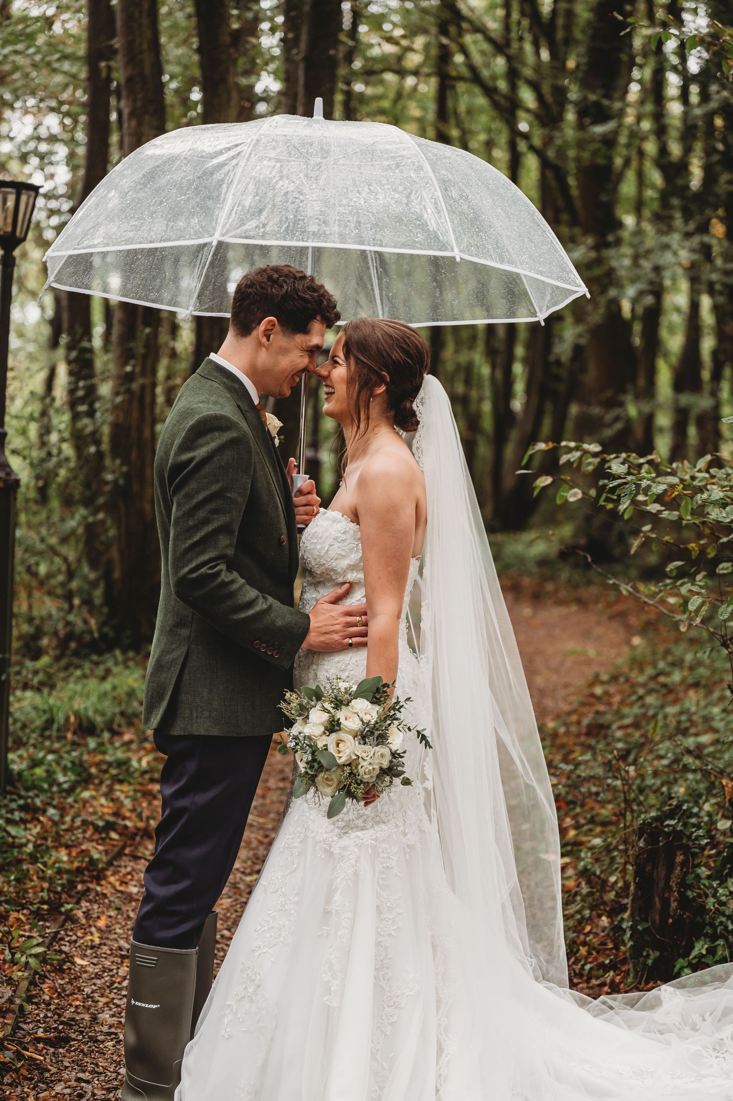 Newlyweds share an intimate moment under a transparent umbrella in a wooded setting with a white bouquet.
