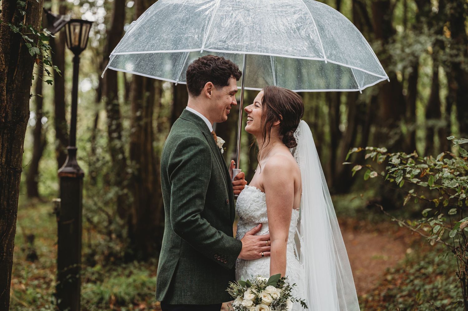 A romantic wedding moment captured under a clear umbrella in a lush forest setting.