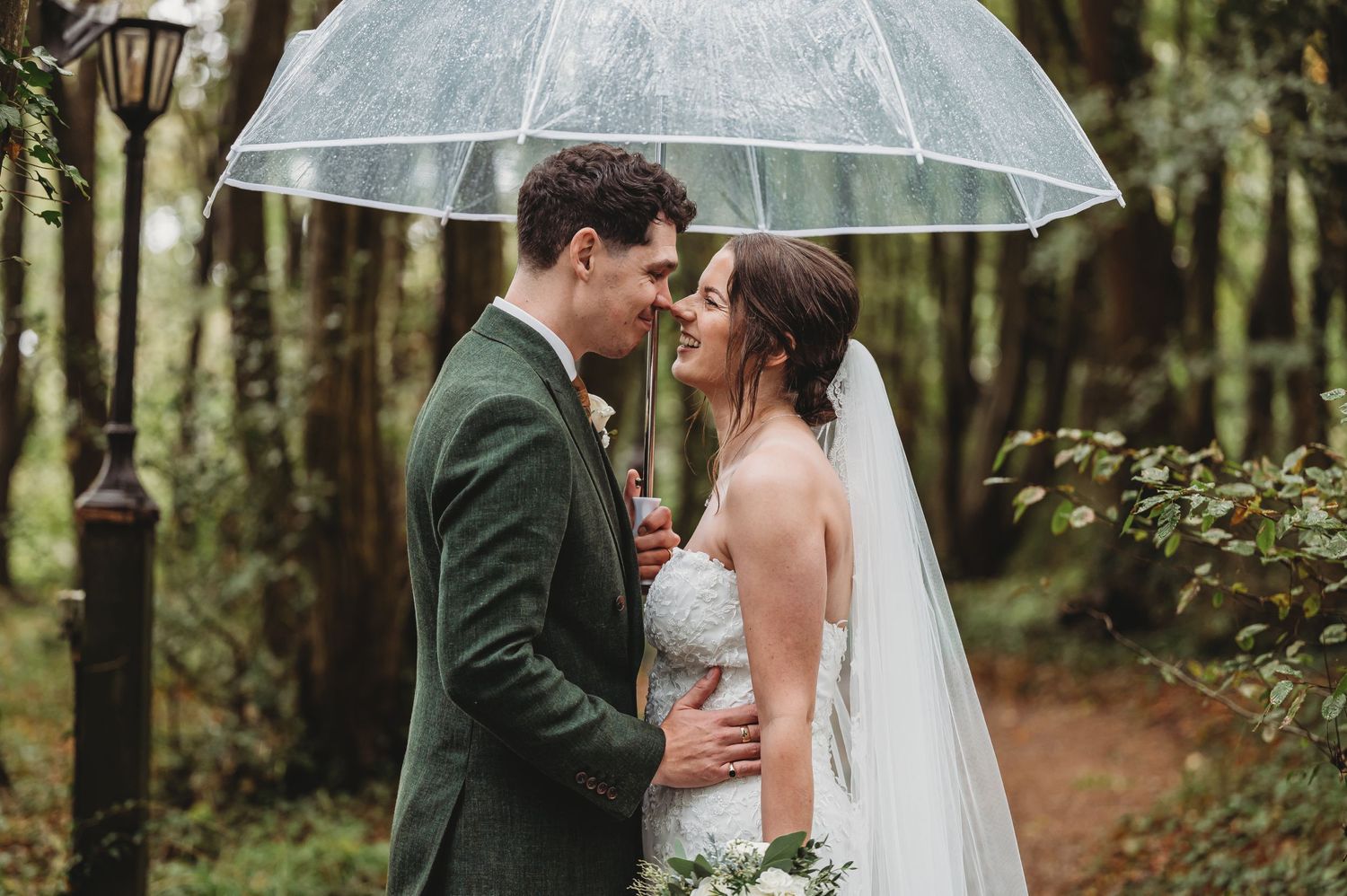 Newlyweds embrace under clear umbrella on wooded forest path during wedding.