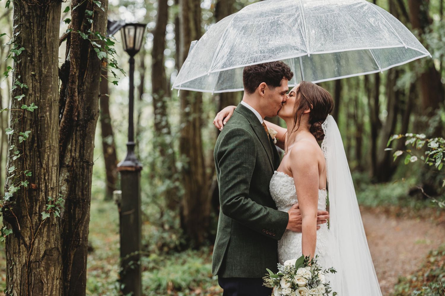 Romantic couple shares intimate moment under clear umbrella in forest wedding setting.