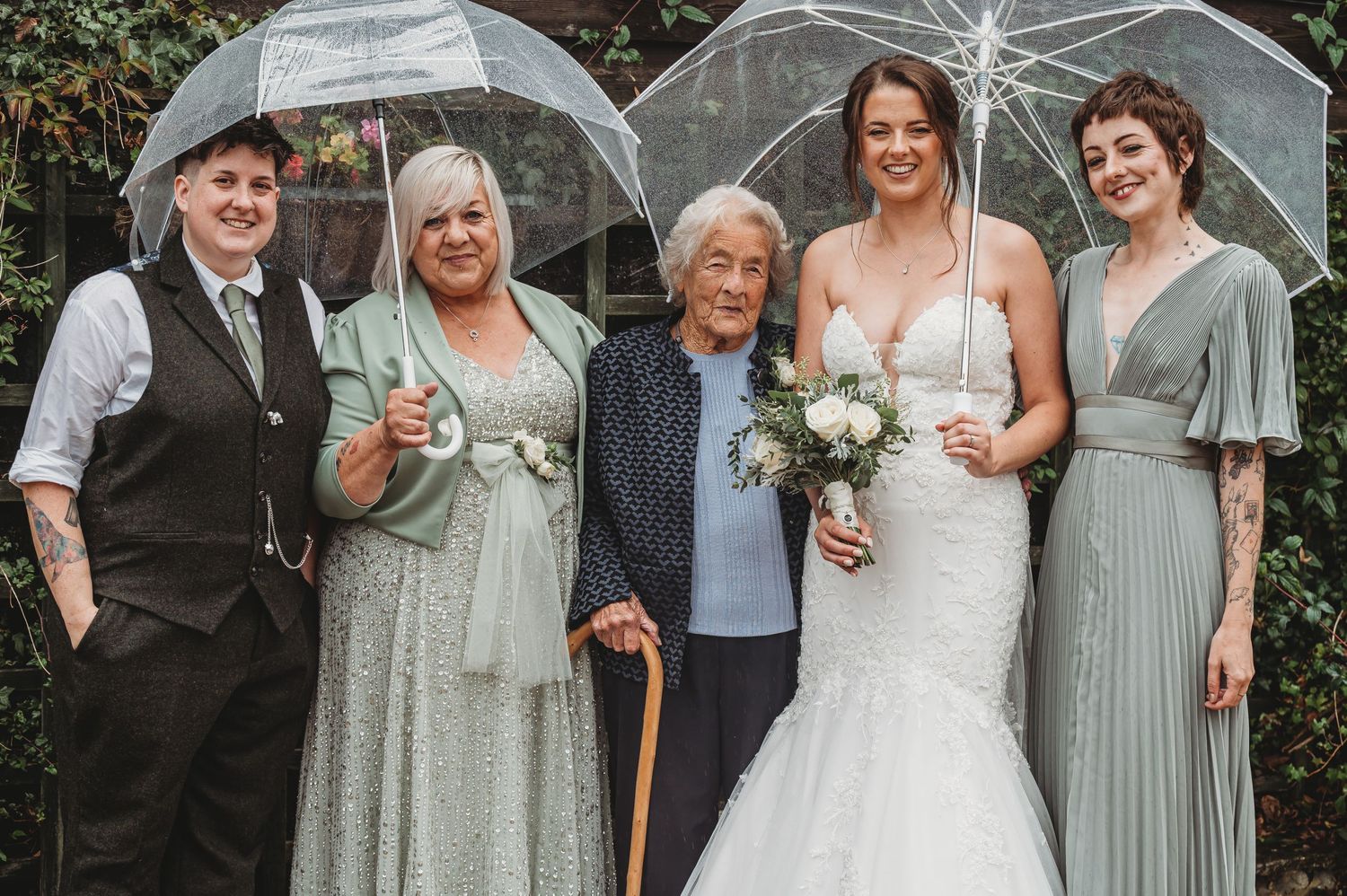Wedding party poses with clear umbrellas while wearing elegant formal attire in shades of sage green and white.