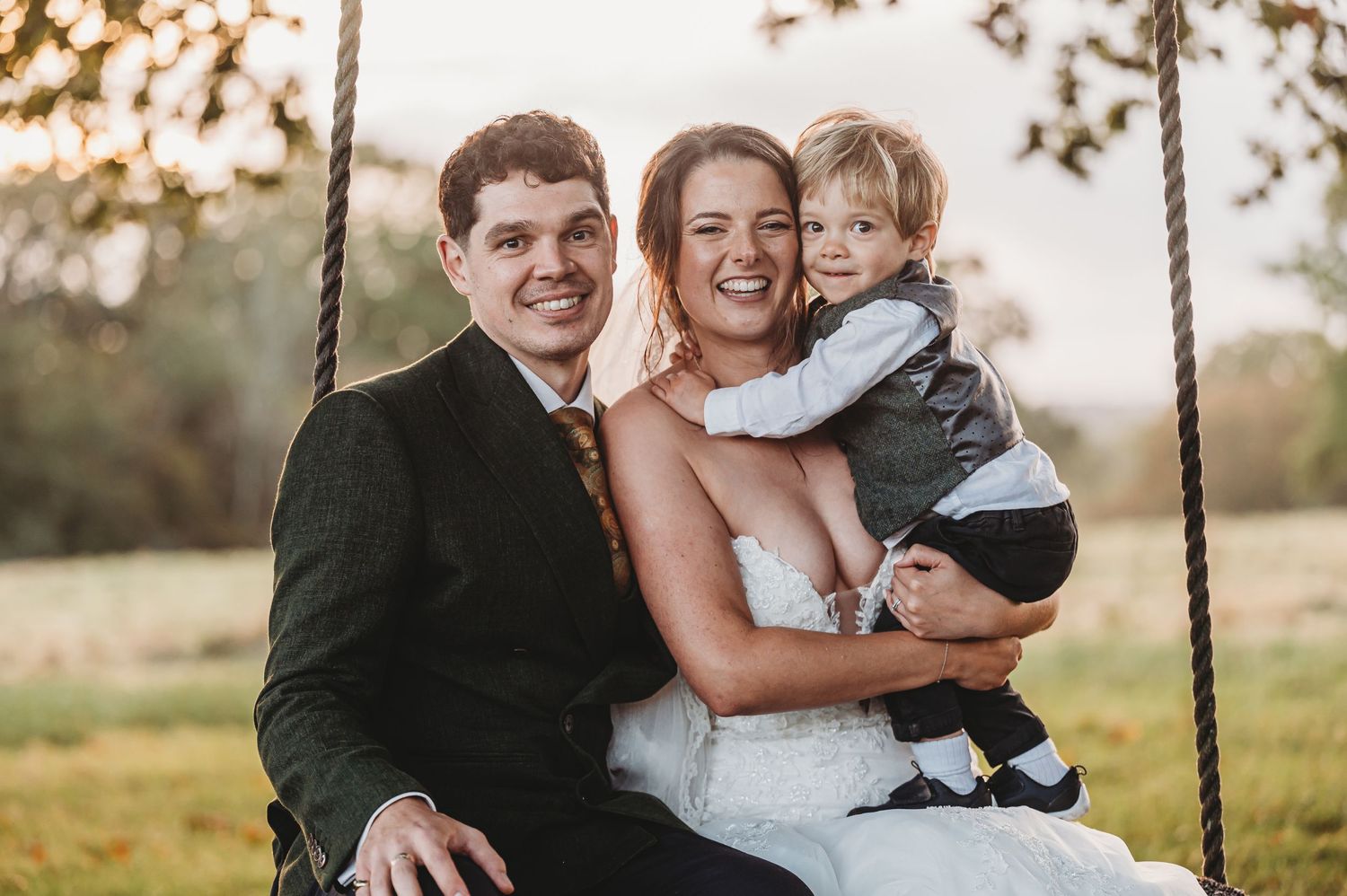 A happy wedding couple sits on a swing at sunset while holding a young child, creating a warm family moment.