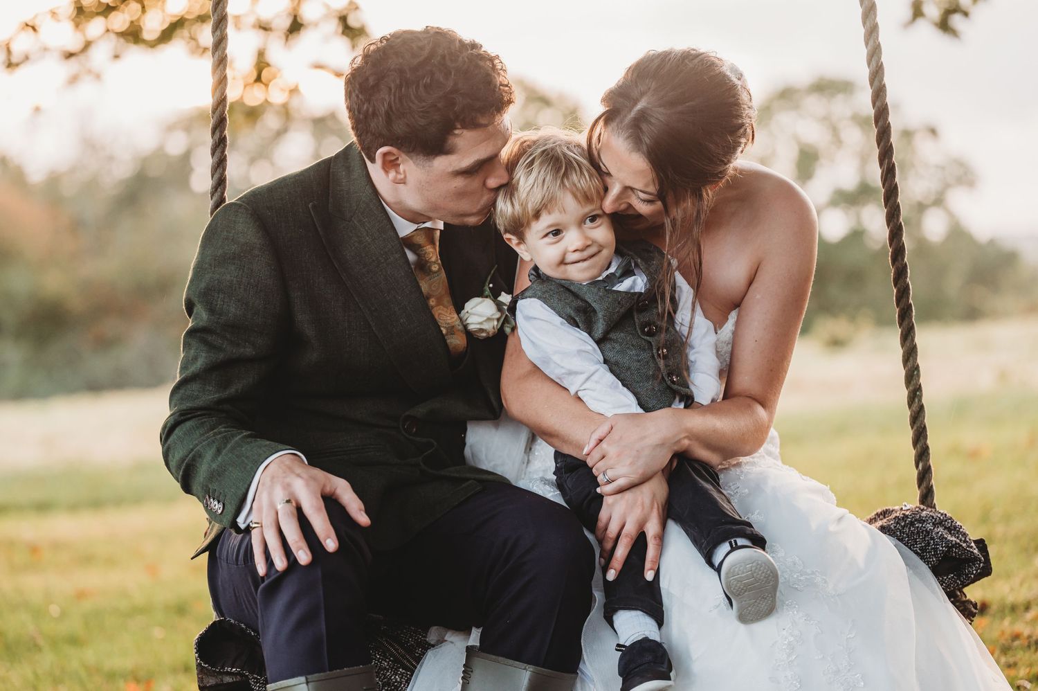 A couple in wedding attire shares a tender moment with their child on a wooden swing during golden hour.