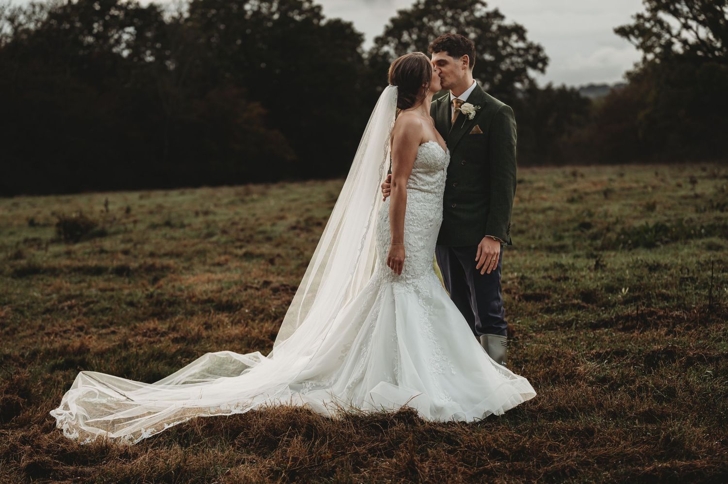 A newly married couple embraces on a rustic field at dusk with a flowing white wedding dress and long veil.