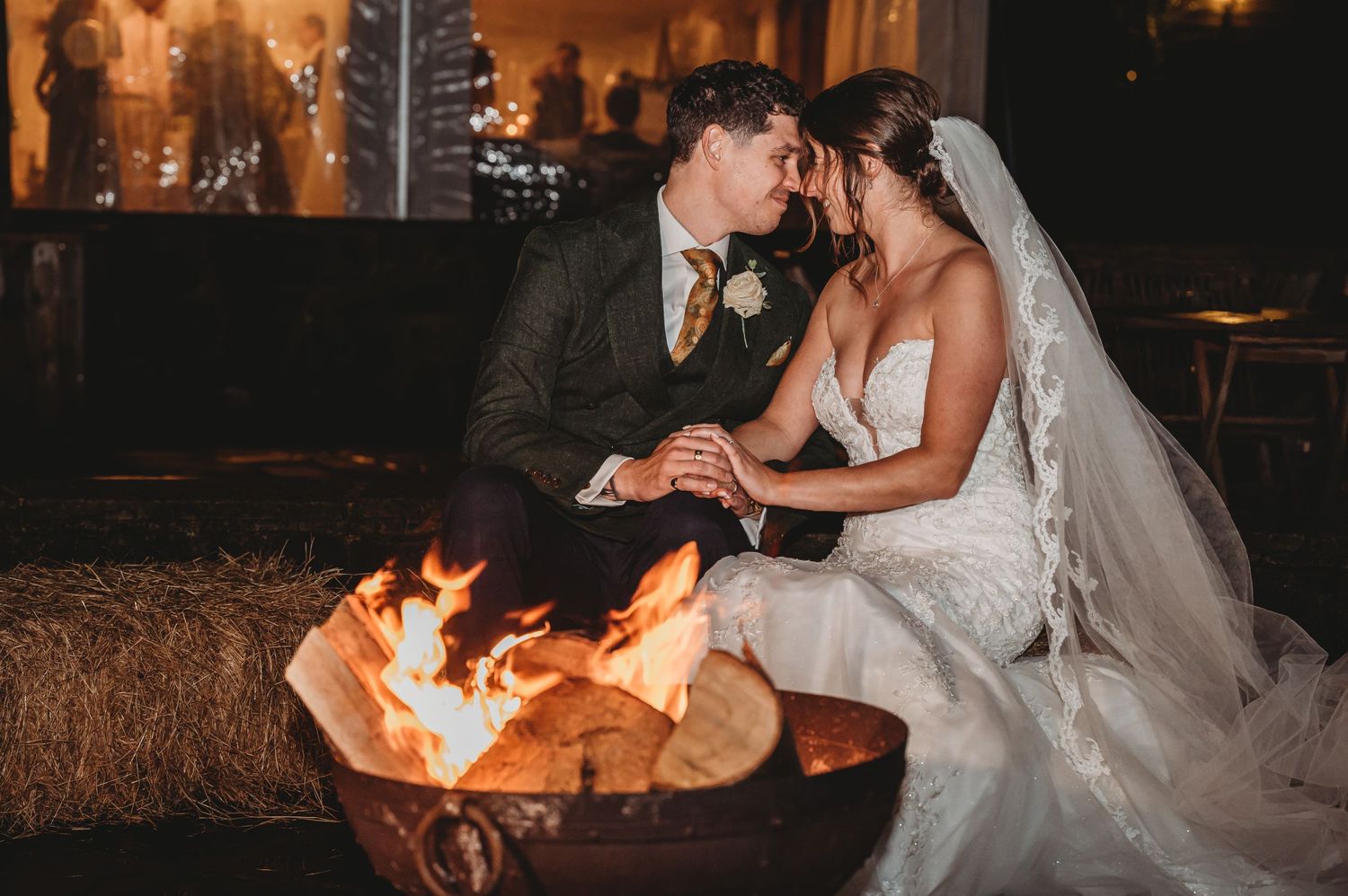 A couple in wedding attire share an intimate moment while sitting beside a glowing fire pit at night.