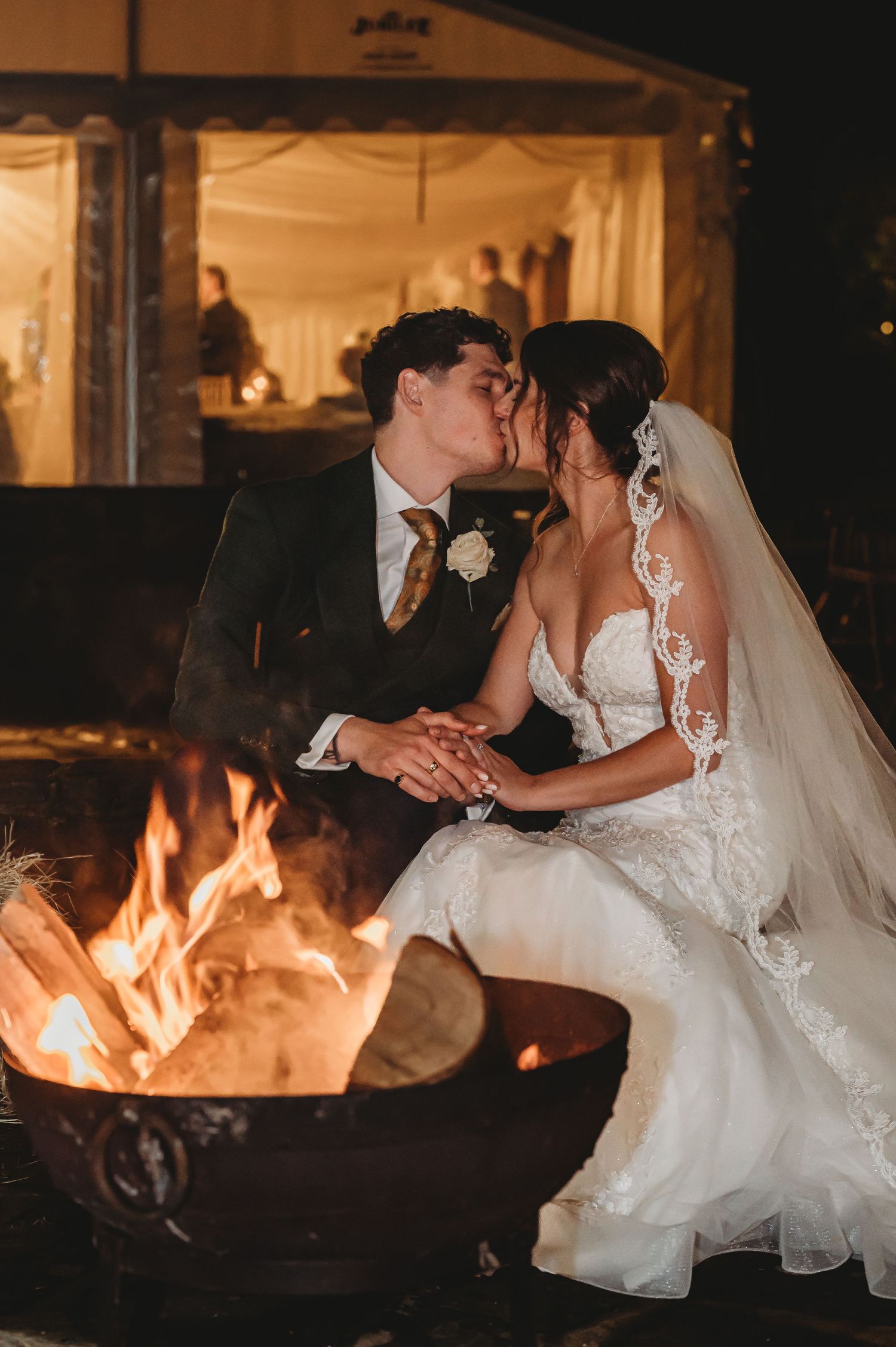 A romantic wedding couple sharing a kiss by a glowing fire pit outdoors, with warm lighting from a nearby tent.