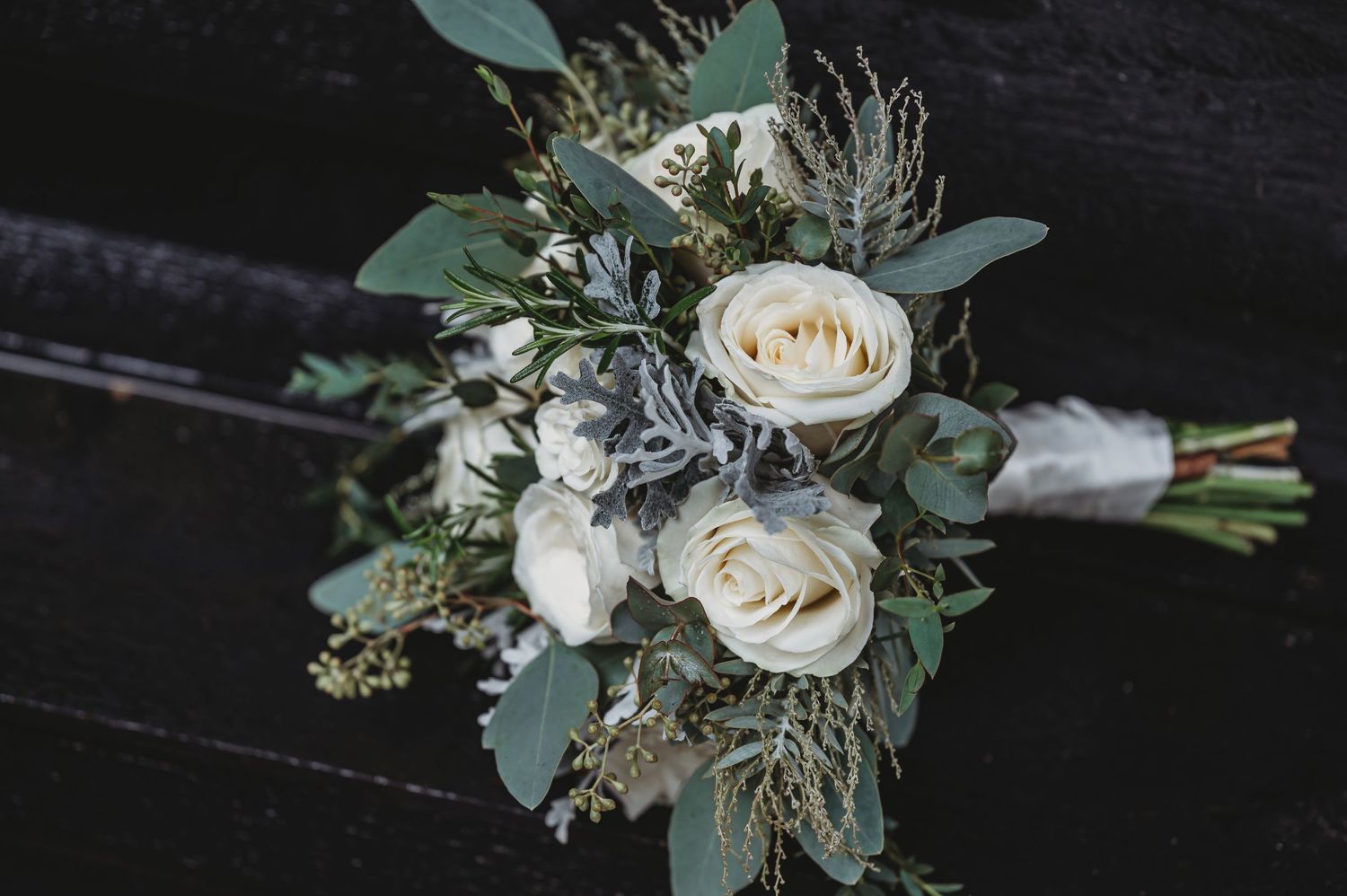 A rustic wedding bouquet with white roses, eucalyptus leaves and silver-toned dried elements tied with ribbon.