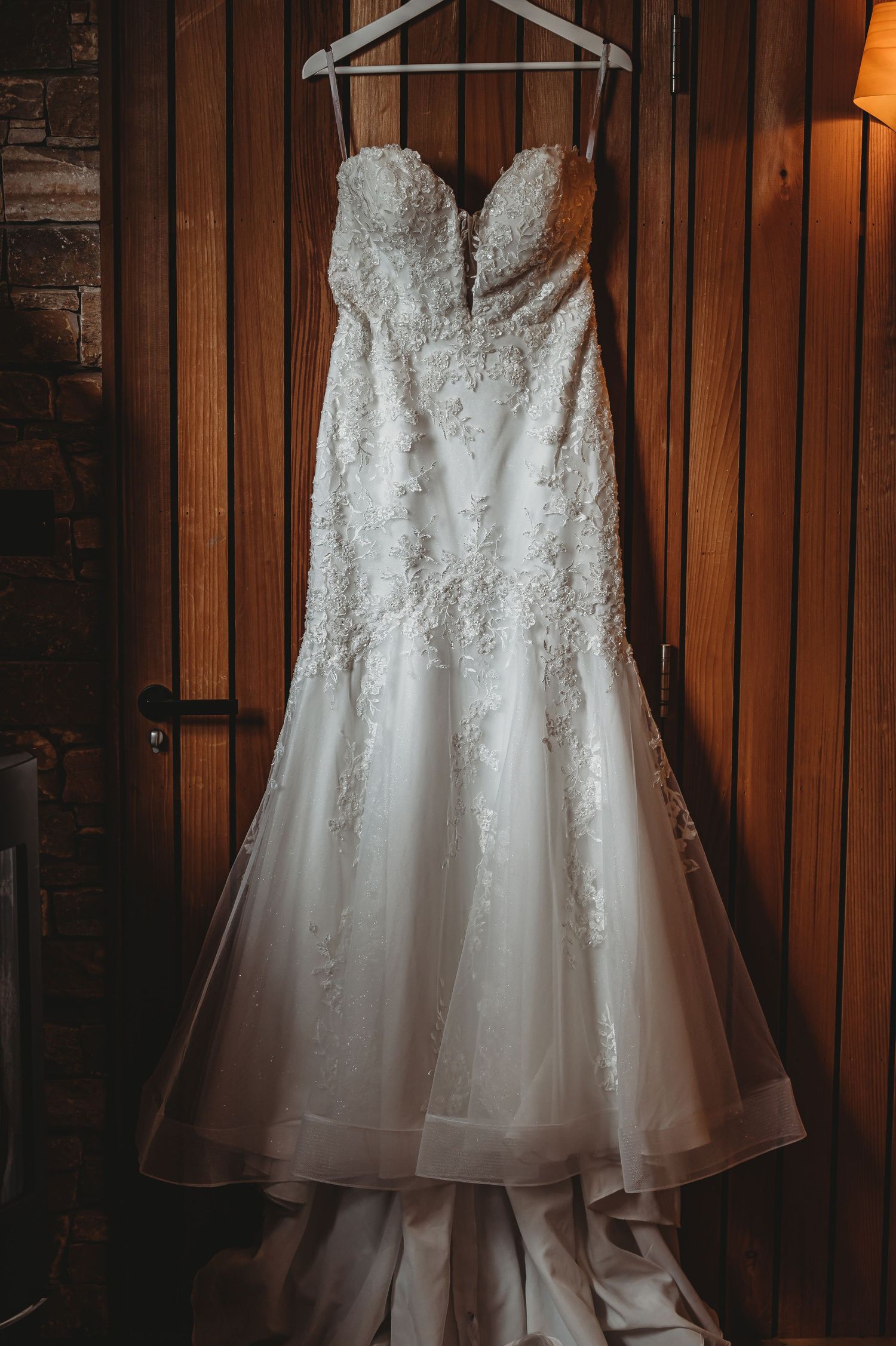 A white lace wedding dress with a sweetheart neckline hangs against wooden paneling.
