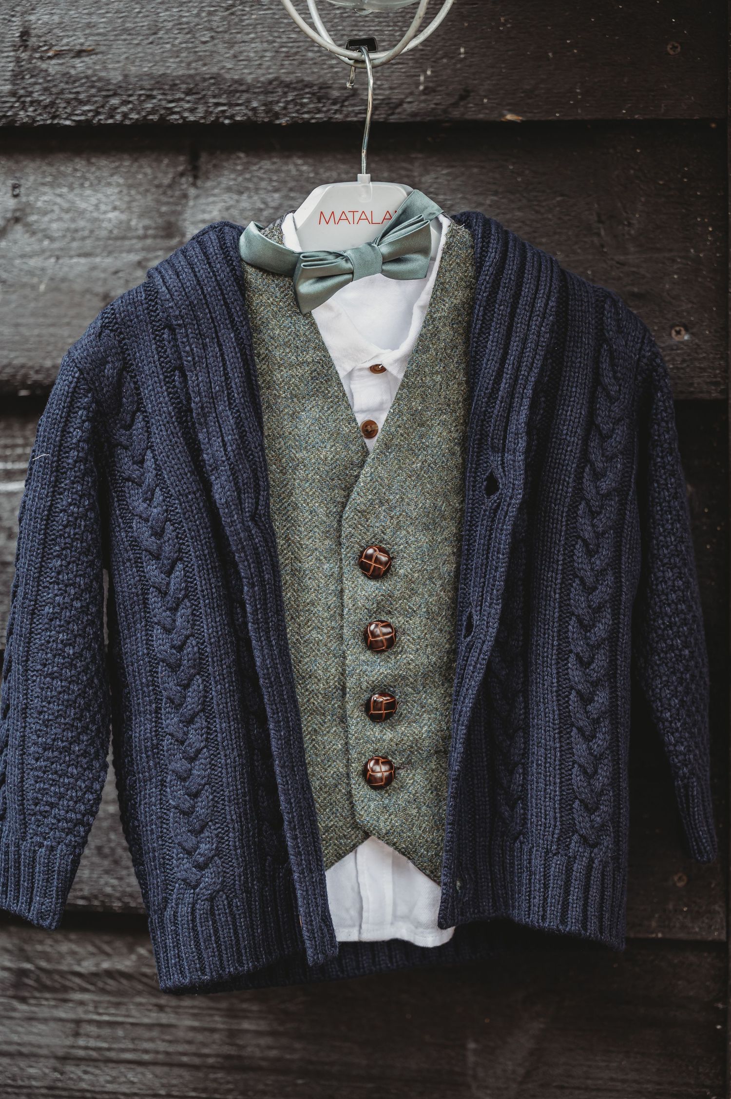 Stylish layered outfit with navy cable knit cardigan, sage green tweed vest, and light bow tie hanging on wooden wall.