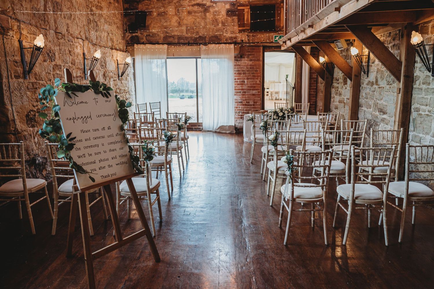 Rustic indoor wedding venue with exposed brick walls, white chairs arranged in rows, and natural light streaming through windows.