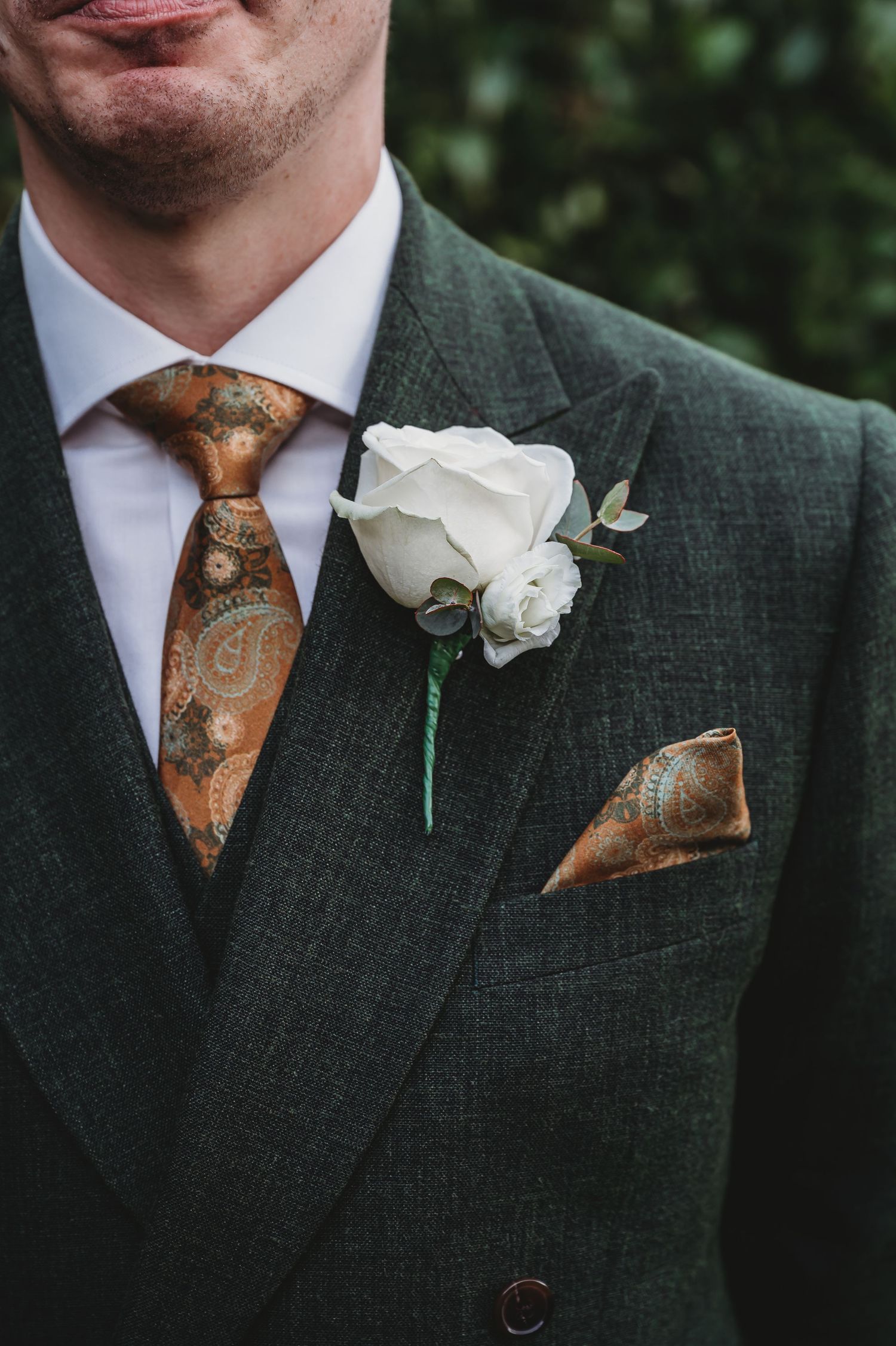 Close-up of a green suit jacket with white rose boutonniere and copper paisley tie.