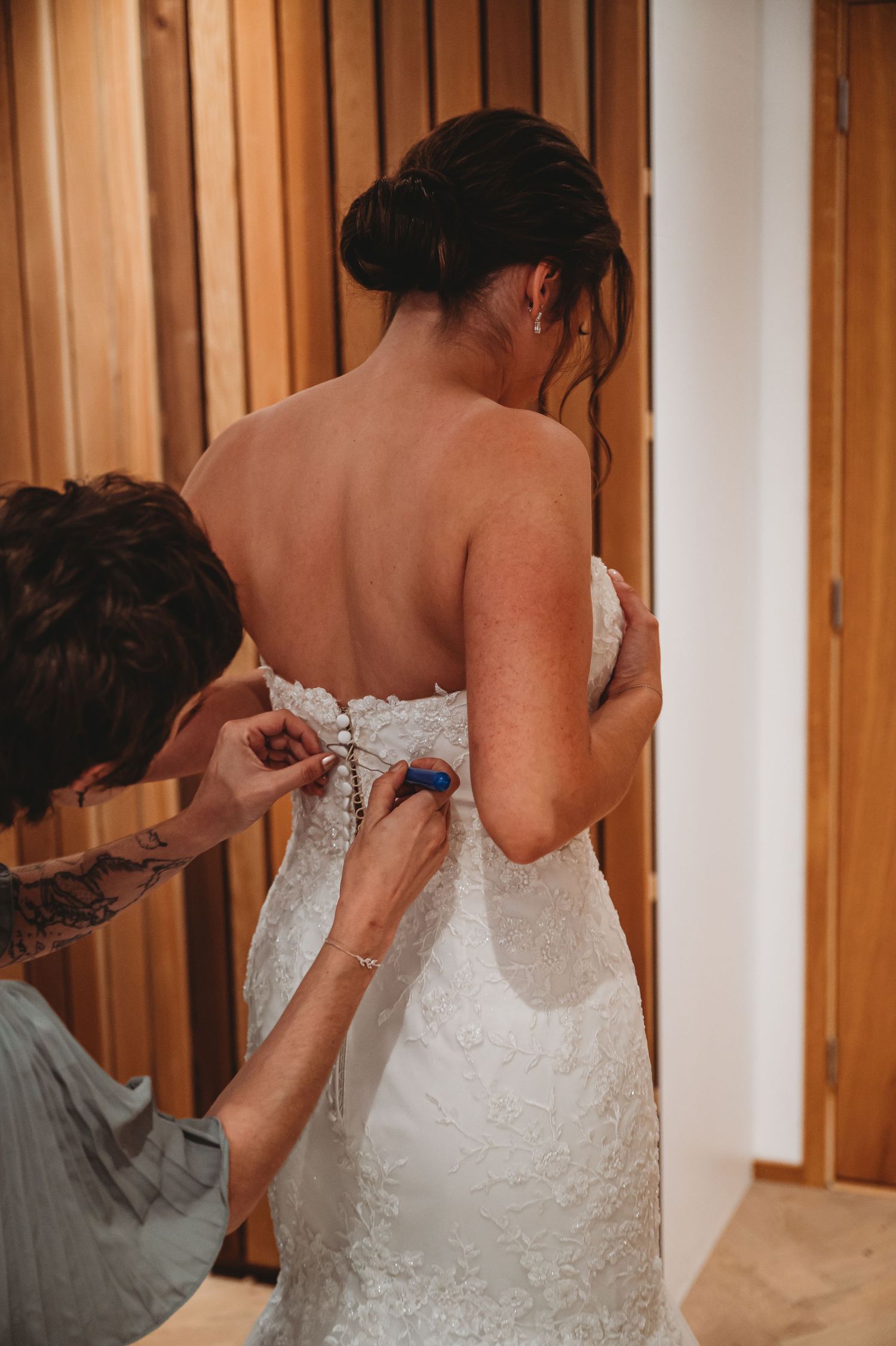 A person helps lace up a white lace wedding dress on a bride getting ready before the ceremony.