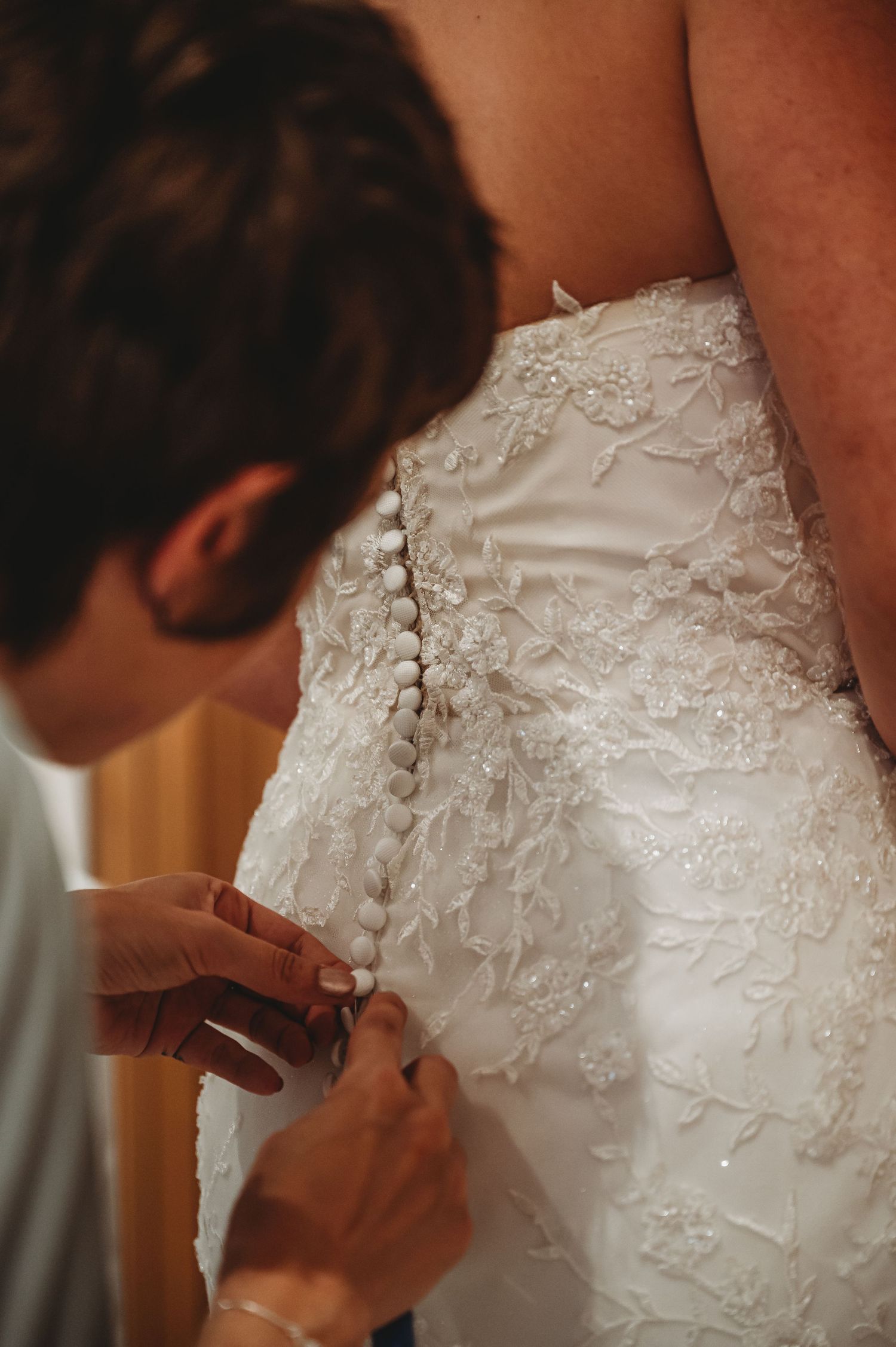Someone helps fasten buttons on an ornate white lace wedding dress with intricate floral embroidery details.