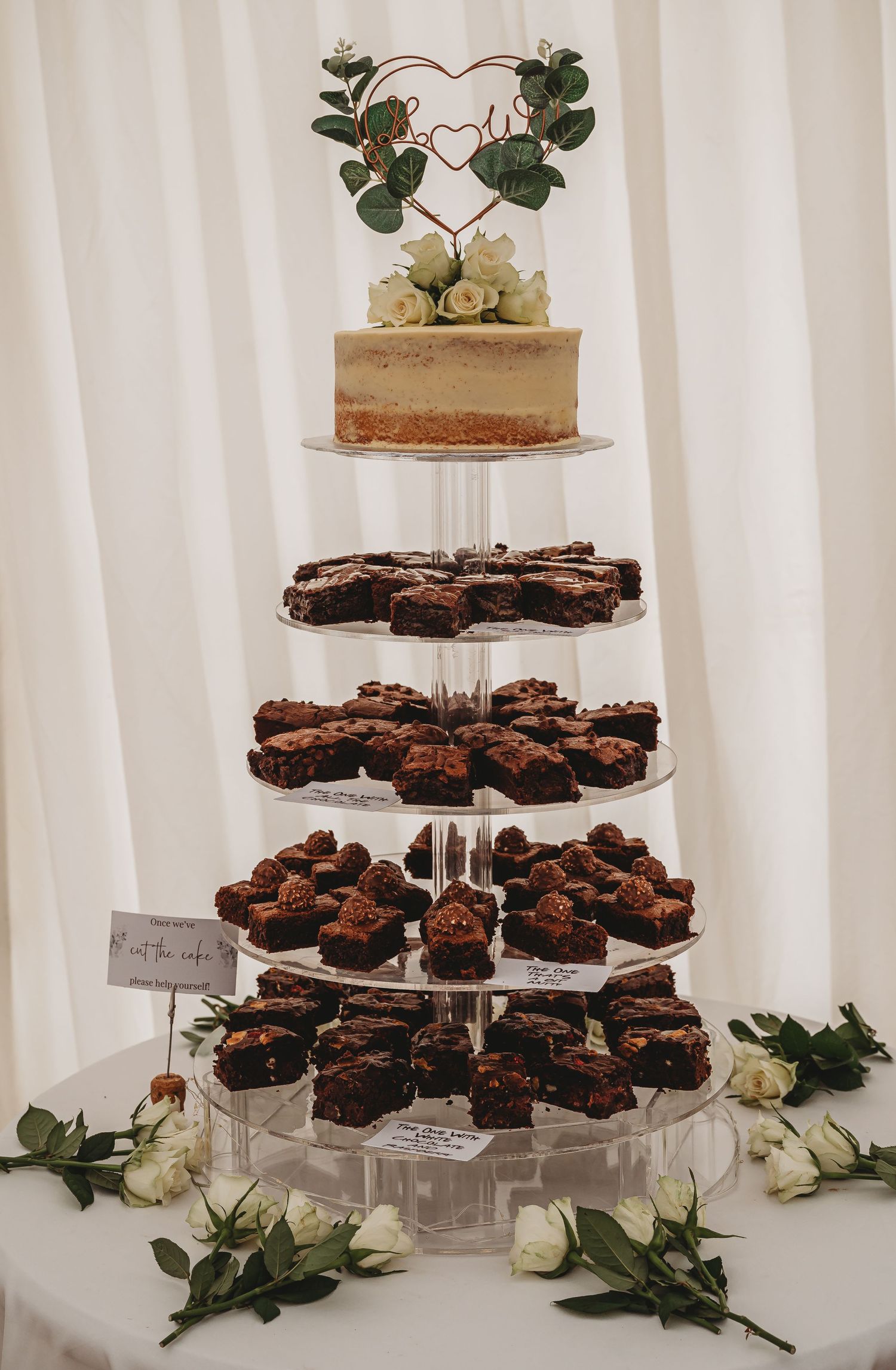 A tiered display of chocolate brownies with a small white cake topper decorated with greenery and roses on a crystal stand.