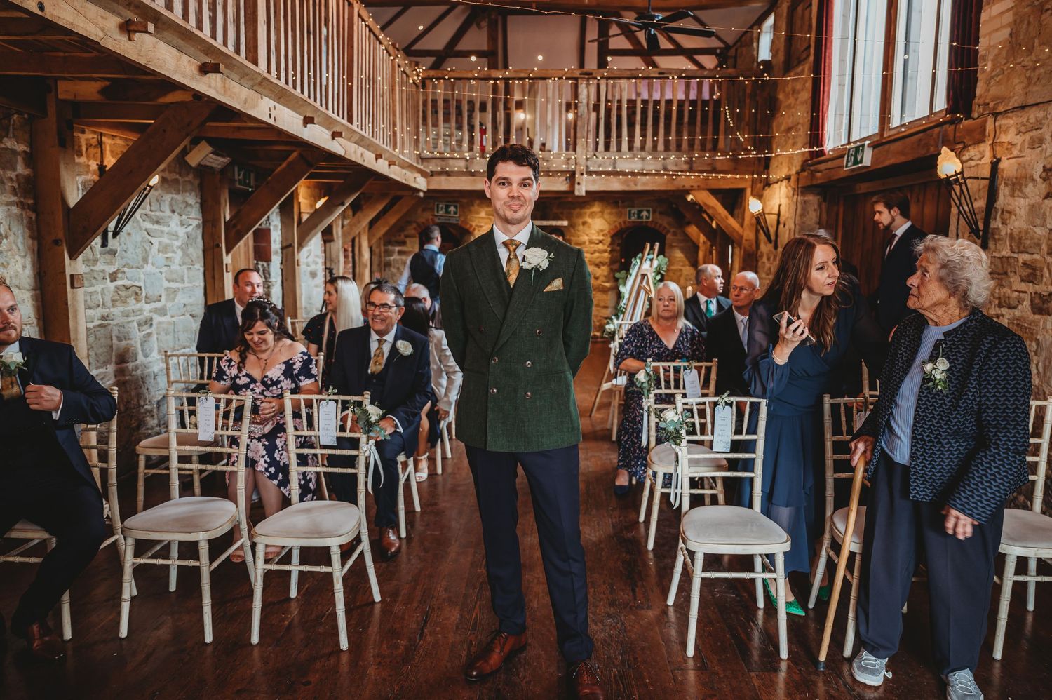 A person in a green suit stands in a rustic barn venue with wooden beams and white chairs lined up for a wedding ceremony.