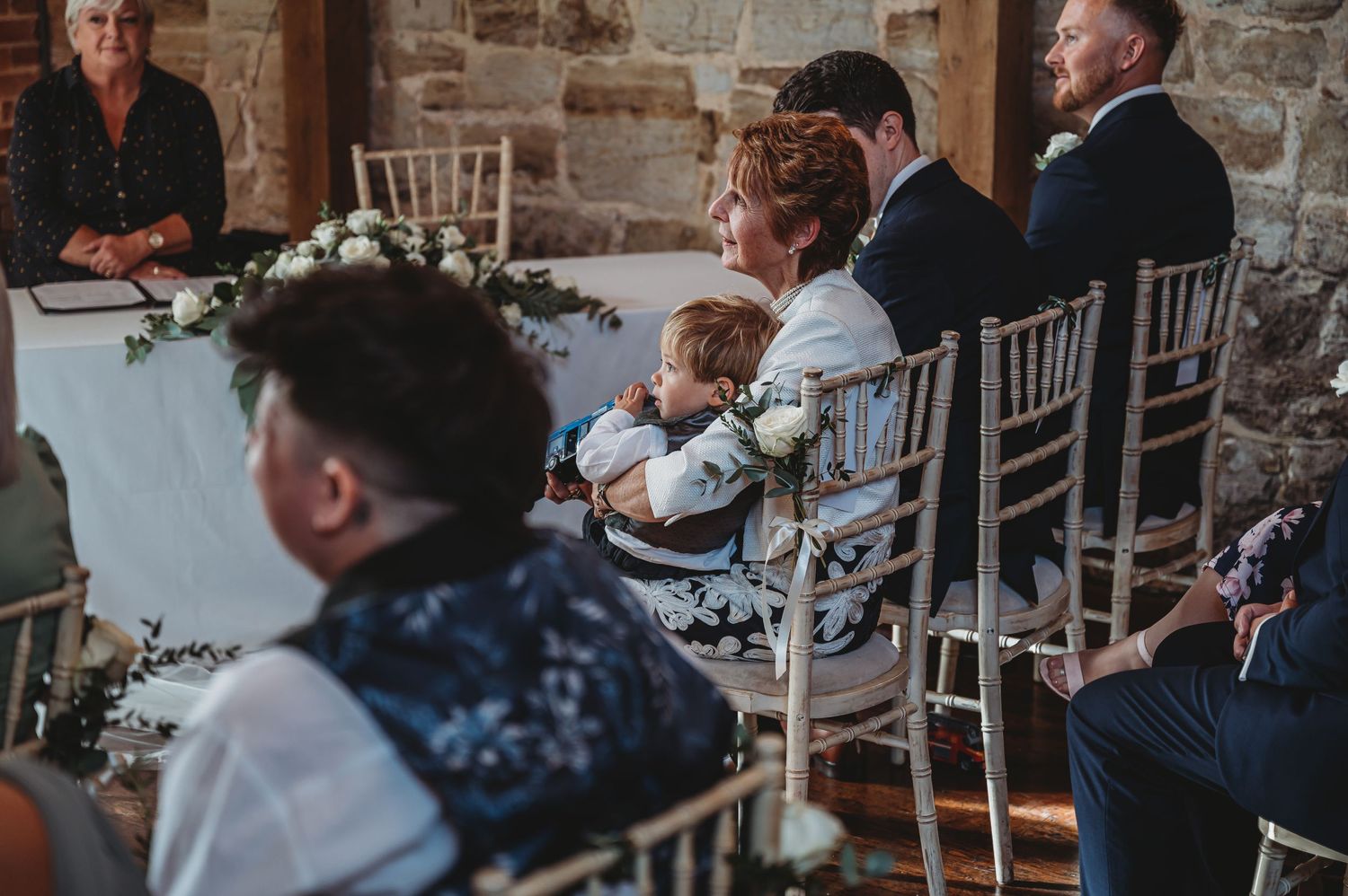Wedding reception guests seated at tables with white chairs in rustic venue.