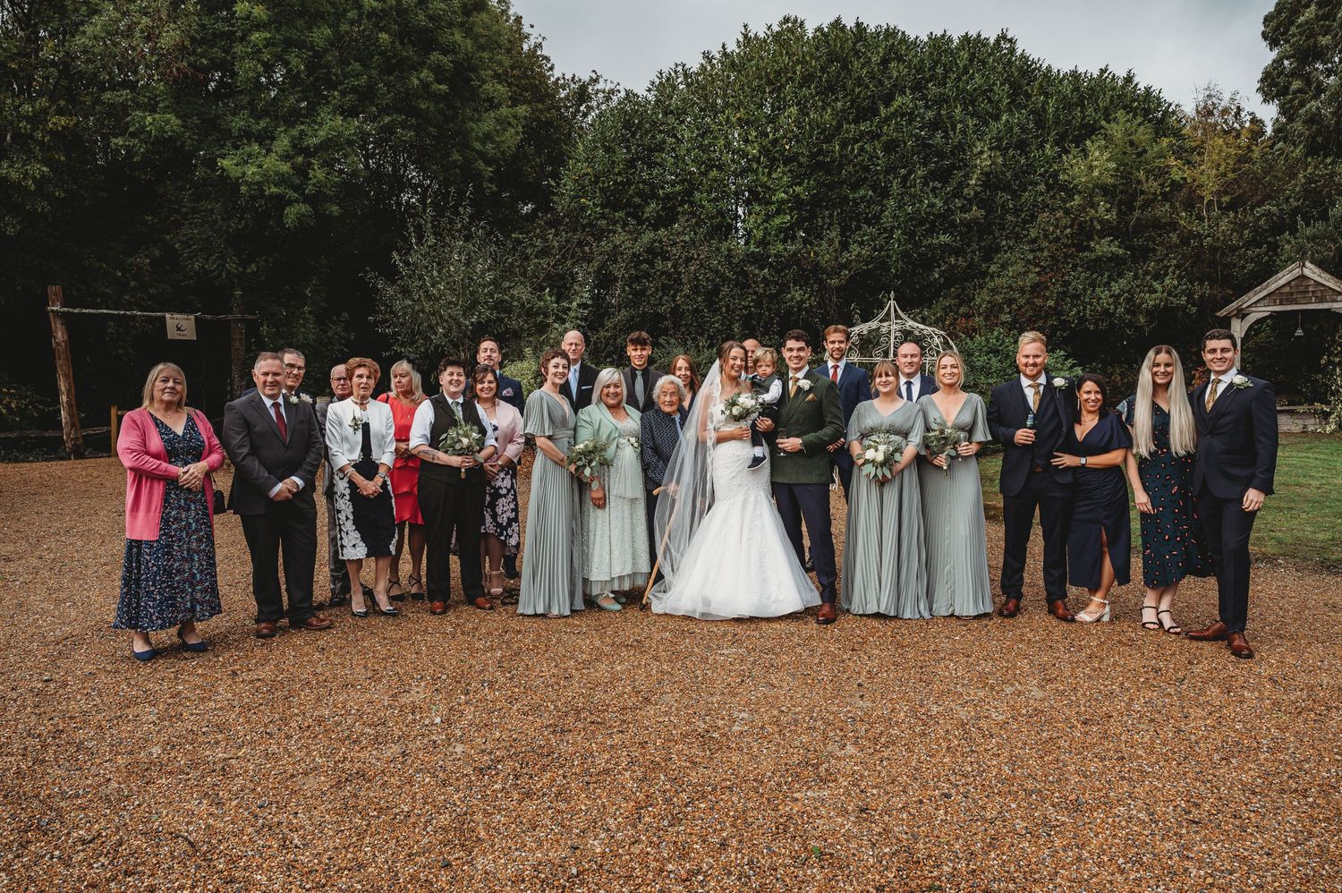 Wedding party and guests lined up outdoors on brown gravel surface with trees in background.