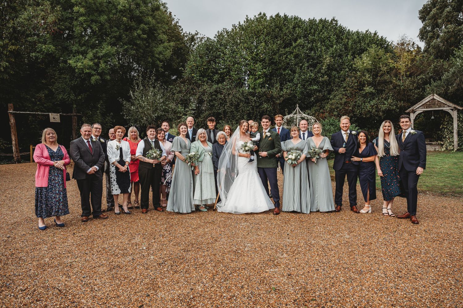 Large wedding party and family group photo outdoors with trees in background.
