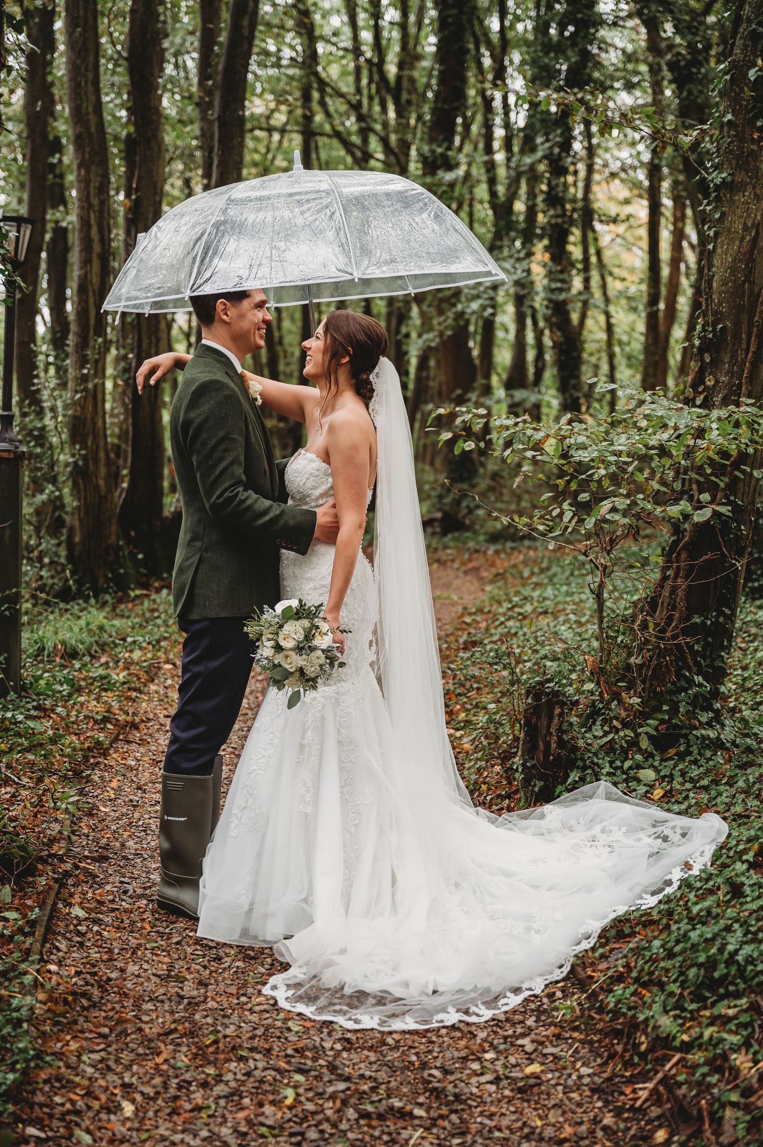 Elegant woodland wedding portrait with couple under clear umbrella on forest path.