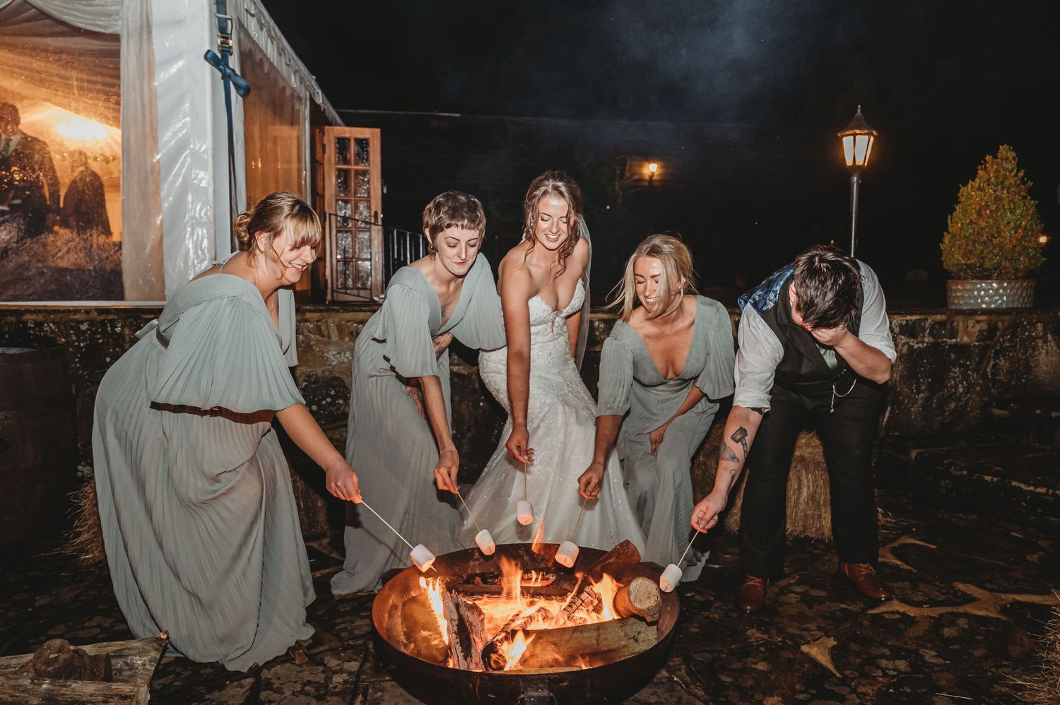 Wedding party members in grey dresses huddle around an outdoor fire pit at night.