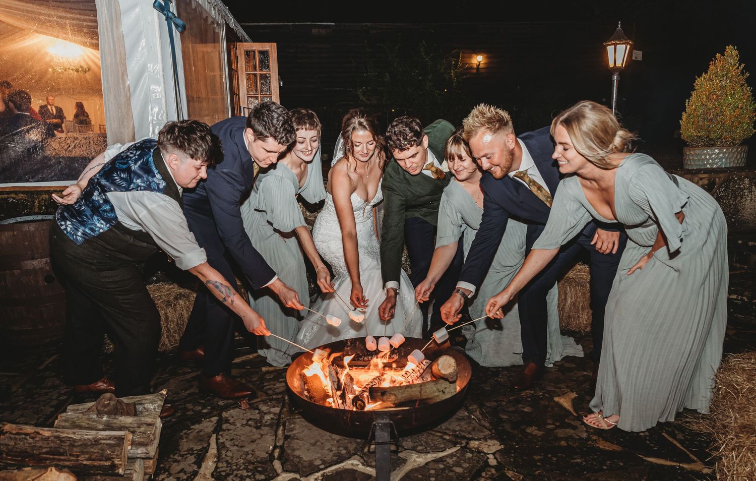 Wedding guests gather around a fire pit at nighttime during outdoor celebration.