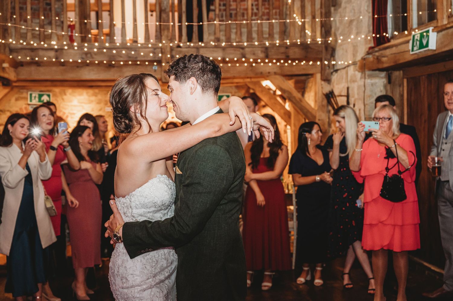 A couple shares their first dance at a wedding reception surrounded by guests in a rustic wooden venue.