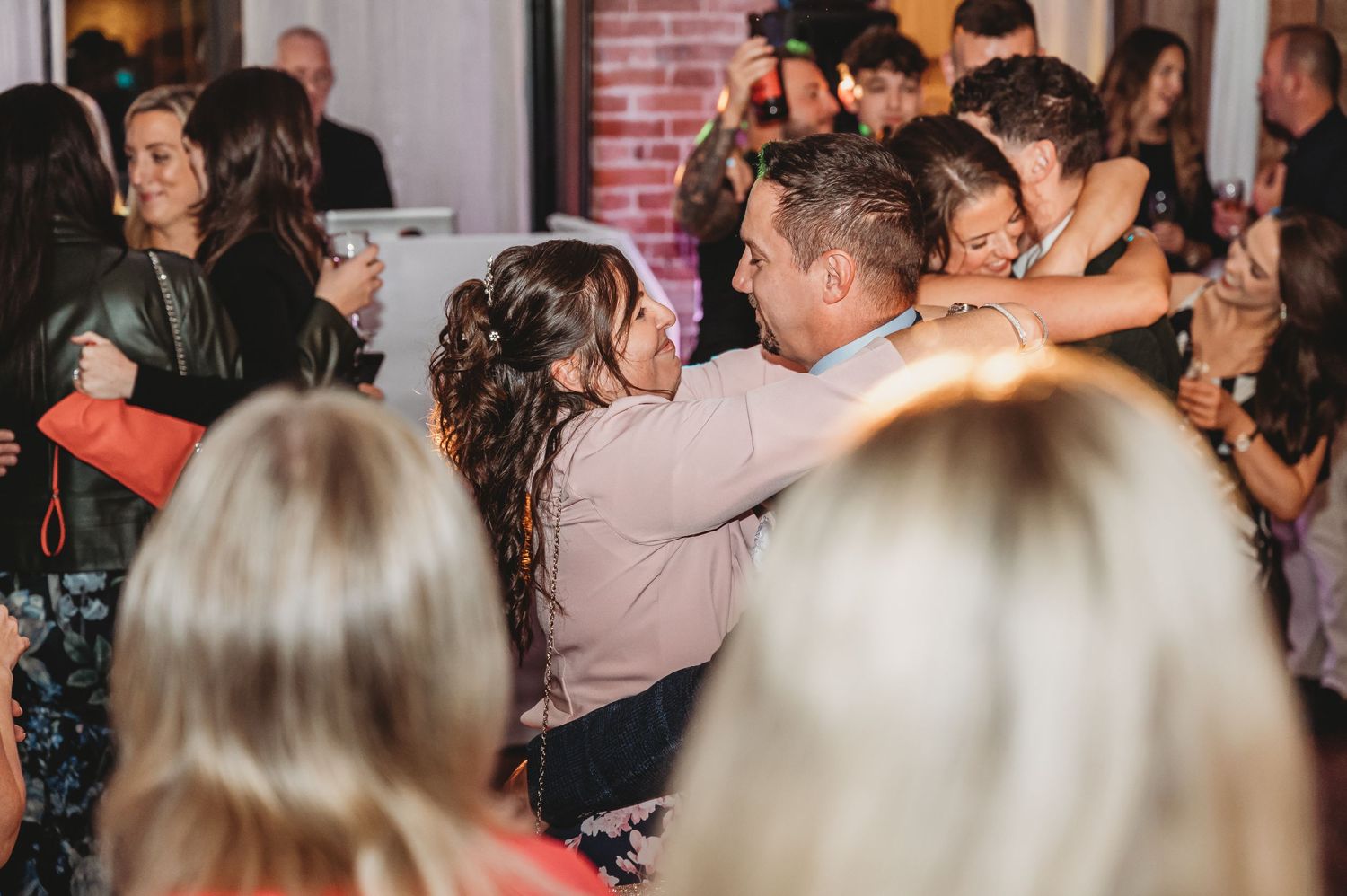 Crowded dance floor at wedding reception with couples dancing under ambient lighting.