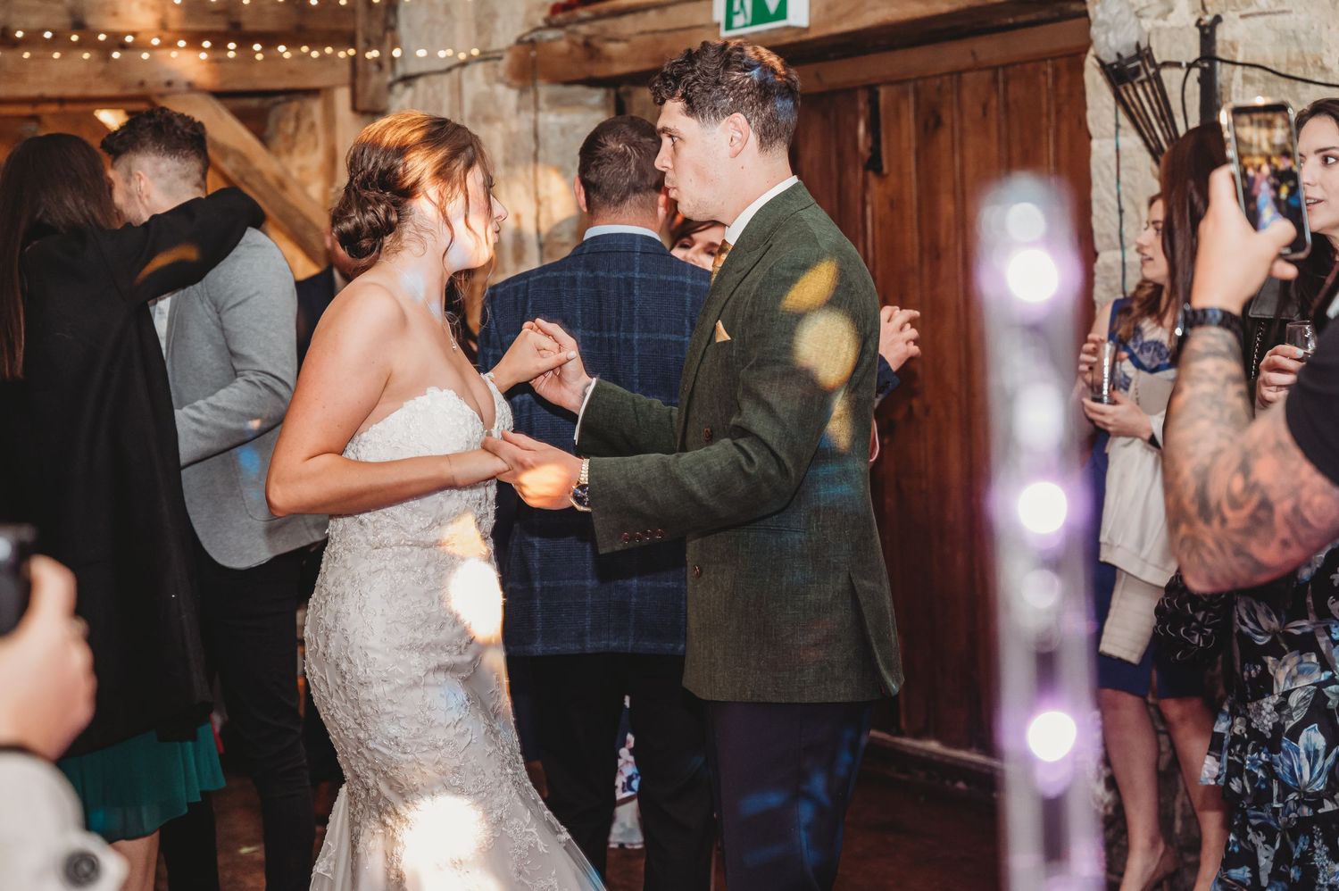 Couples dancing at wedding reception in rustic wooden venue with string lights overhead.