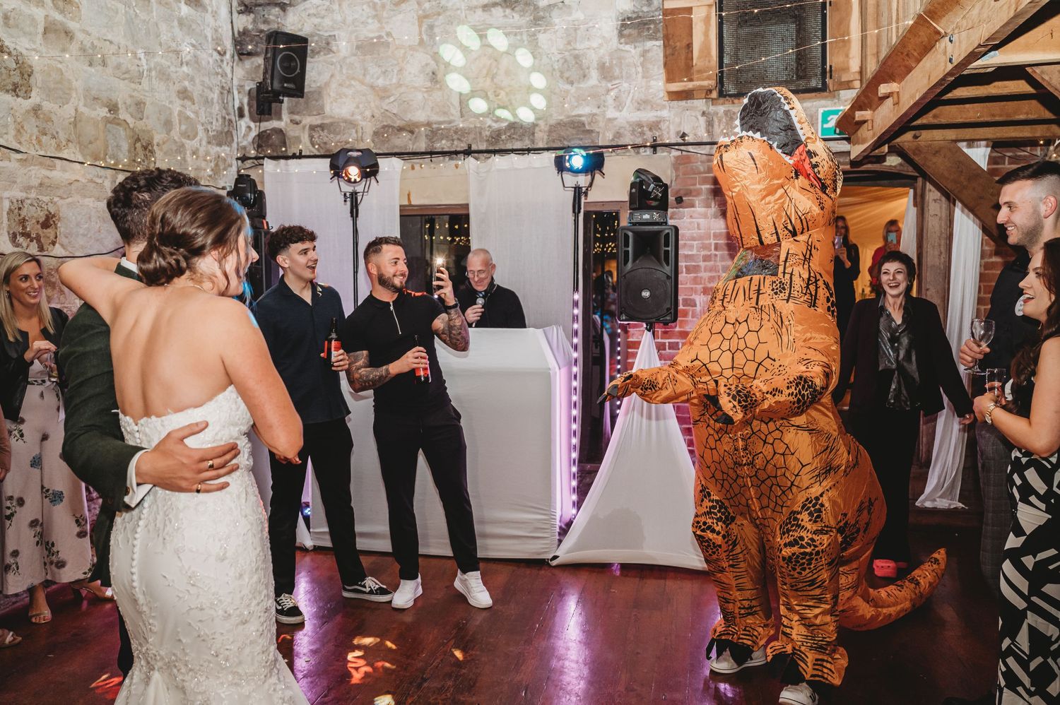 Wedding celebration featuring dance between bride and person in T-Rex costume surrounded by guests.