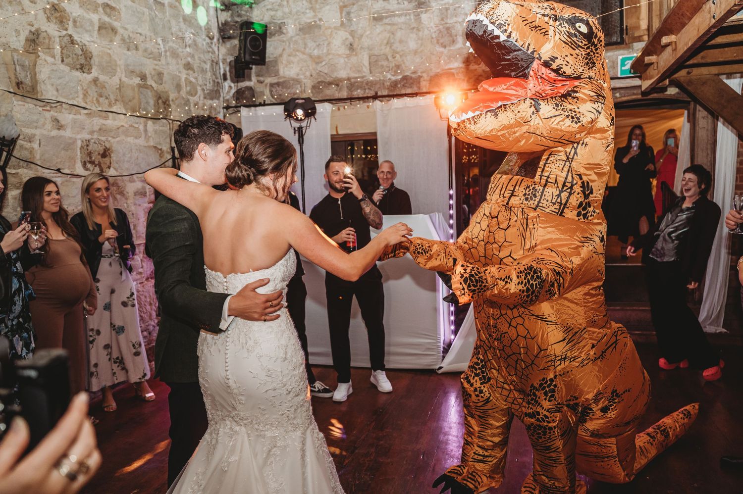 Newlywed in white dress dances with inflatable dinosaur costume at stone-walled reception venue.