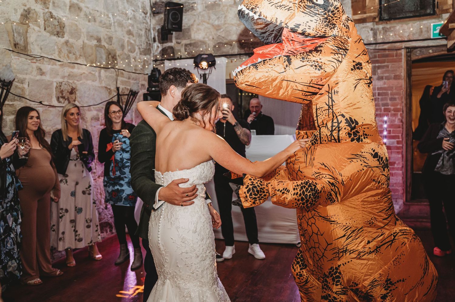 Wedding reception scene with someone in wedding dress dancing with person in inflatable T-Rex costume.