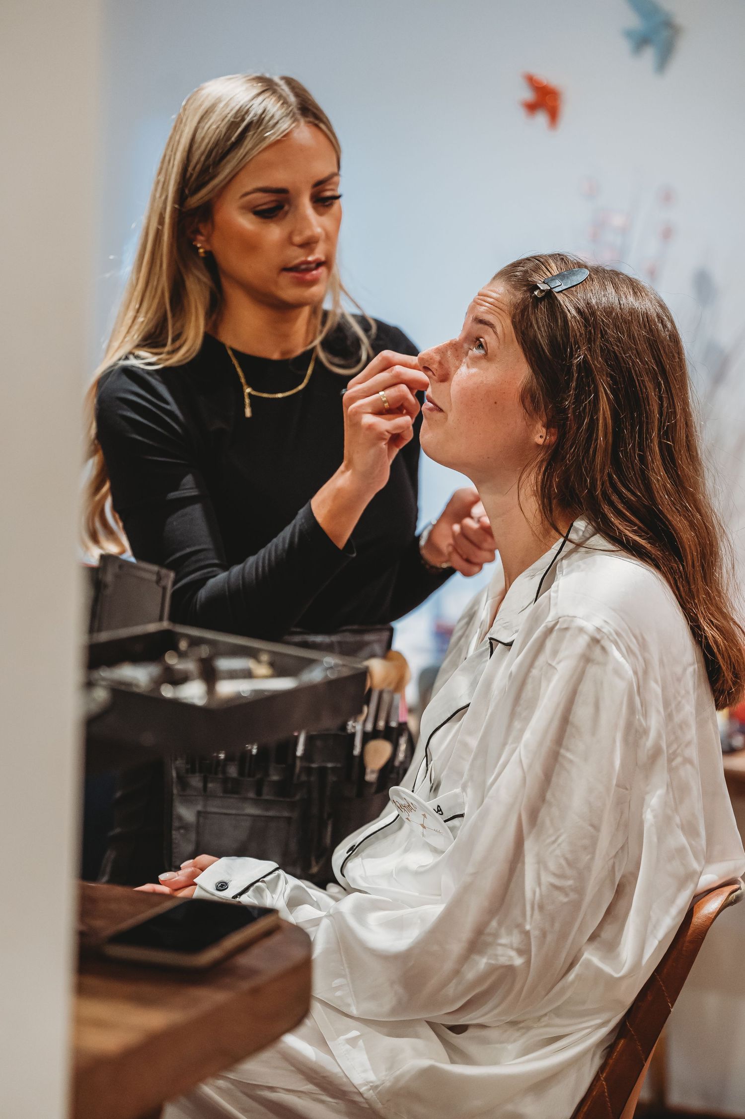 Professional makeup artist applying cosmetics to client wearing white robe in preparation room.