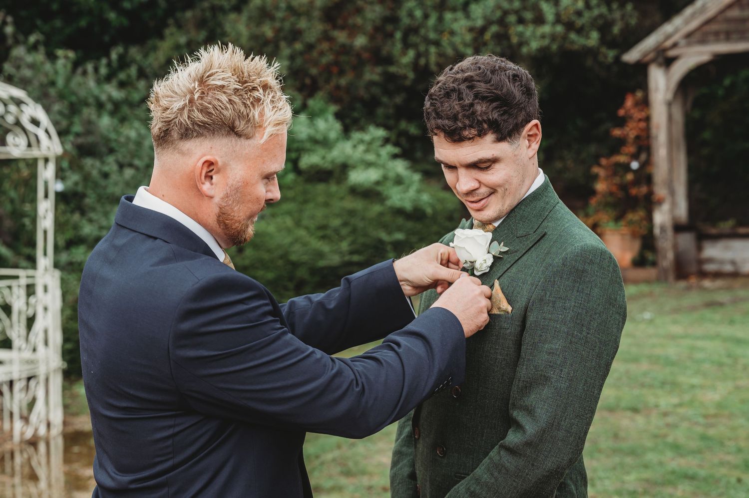 Two people sharing a candid moment while adjusting formal wear in an outdoor garden setting.