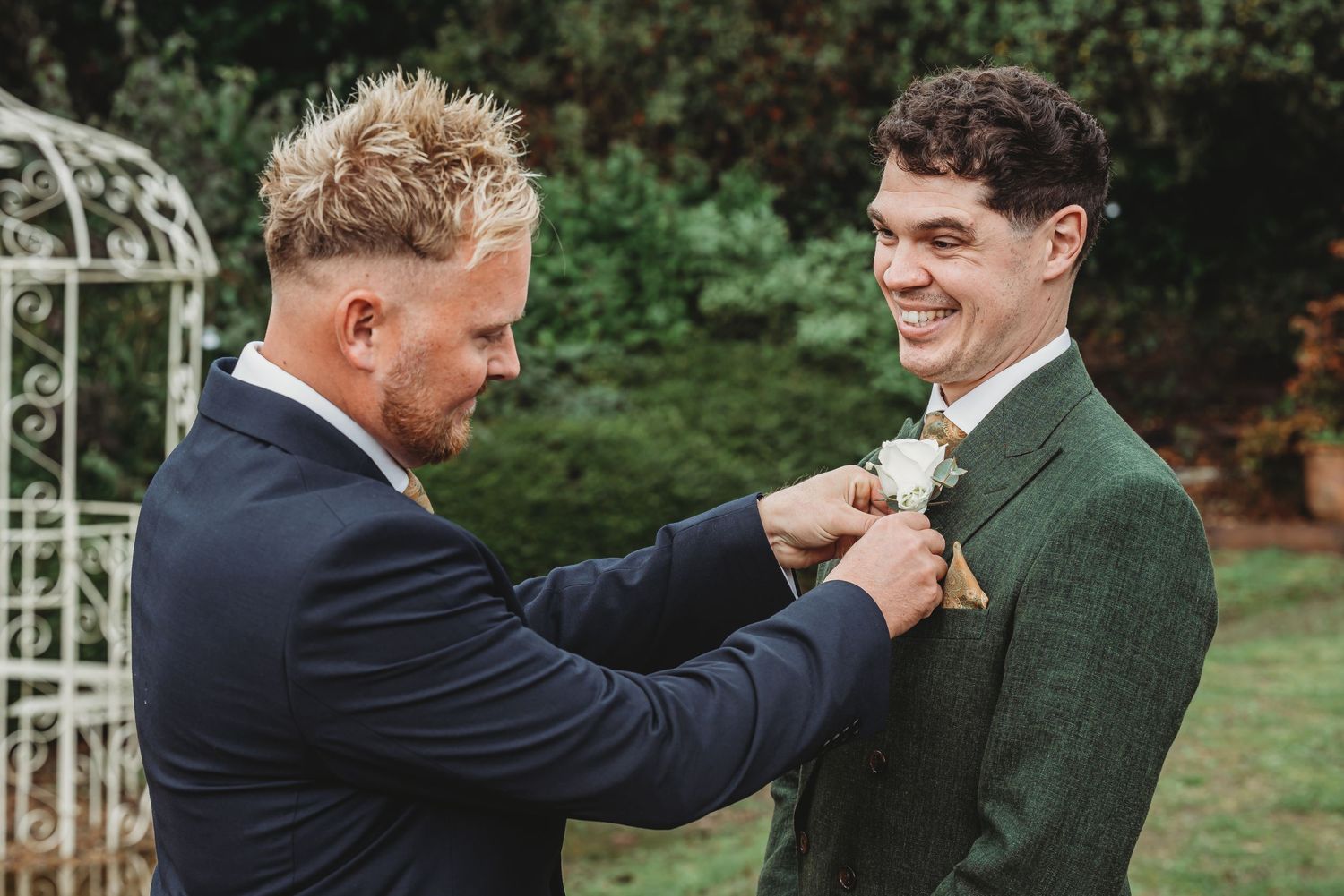 Two people in contrasting suits helping each other with boutonnieres in front of a white garden arch.