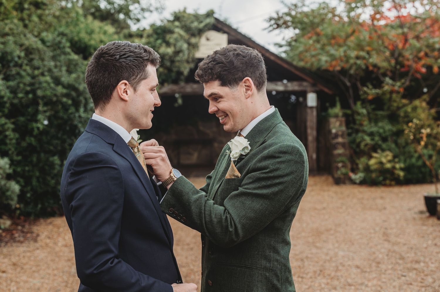 Two people in formal attire sharing a touching moment while adjusting each other's boutonnieres in a garden setting.