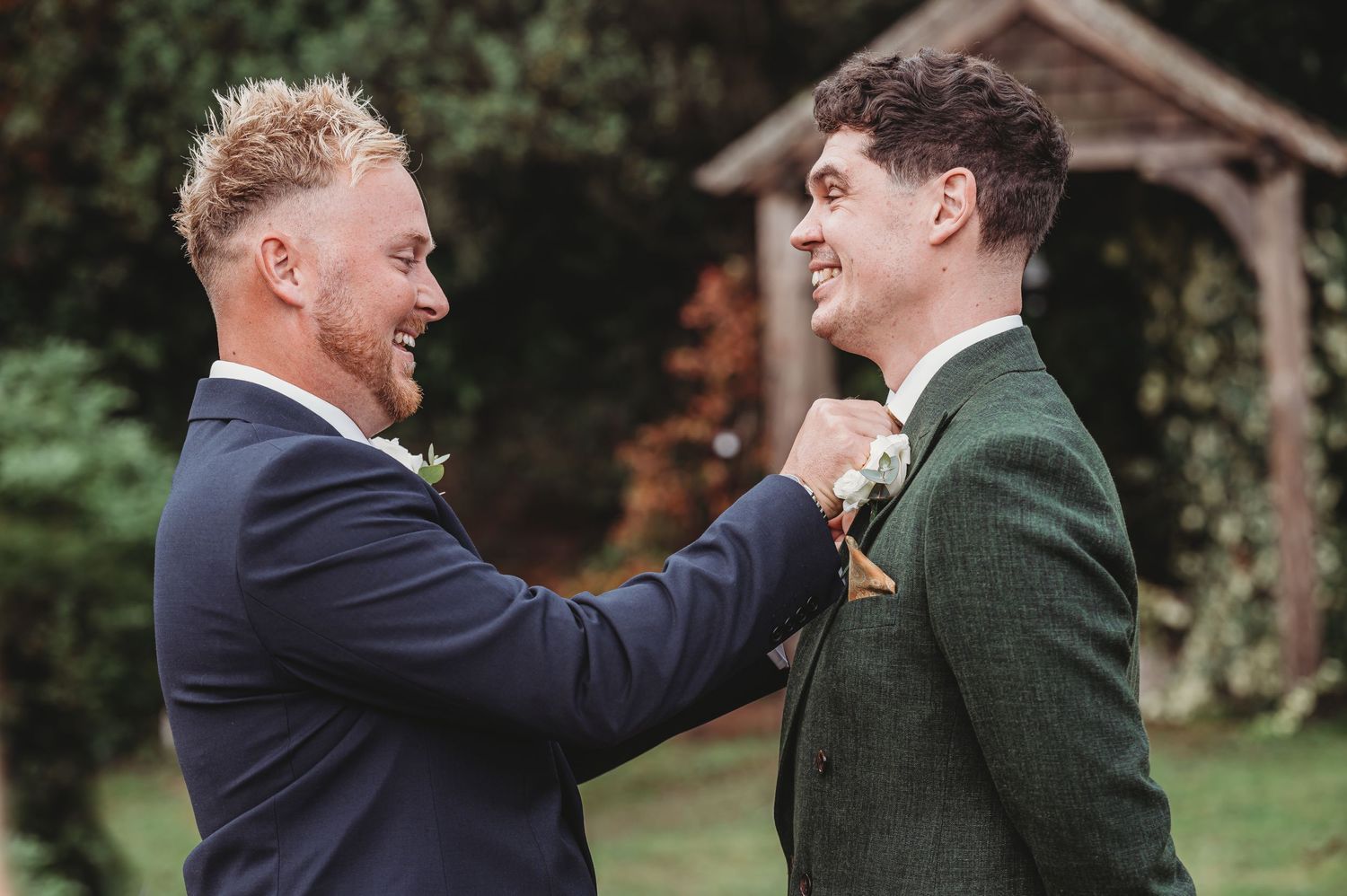Groomsman helps adjust another's boutonniere while sharing a laugh outdoors.
