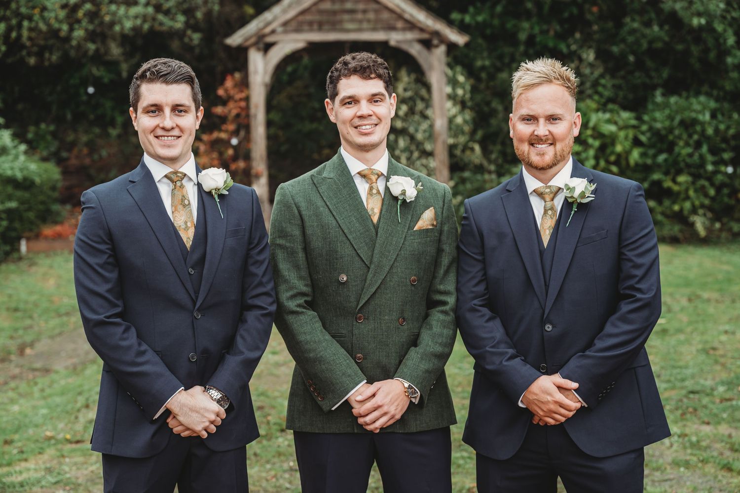 Three groomsmen in coordinating suits pose formally in front of a garden gazebo.