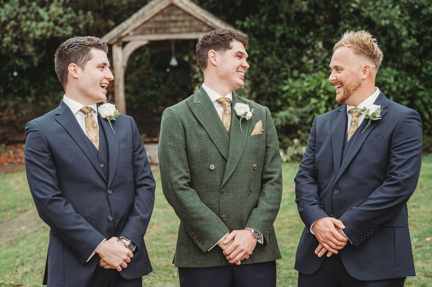 Three groomsmen share a laugh while standing in front of a garden archway.