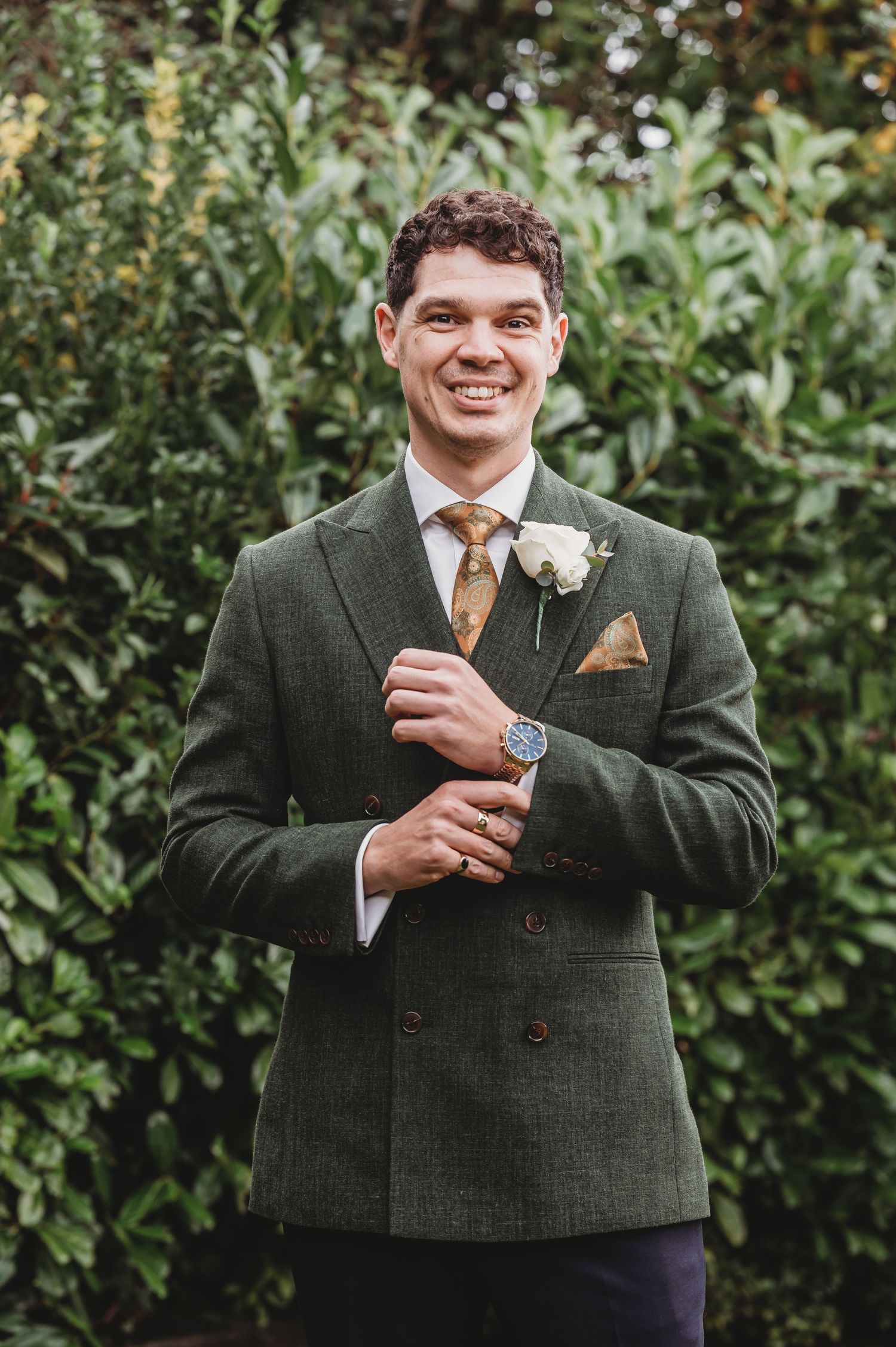 Portrait of groom in tailored green suit with boutonniere against lush foliage.