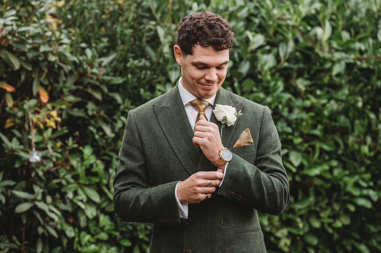 Groom in dark green suit adjusts cufflinks against garden backdrop.