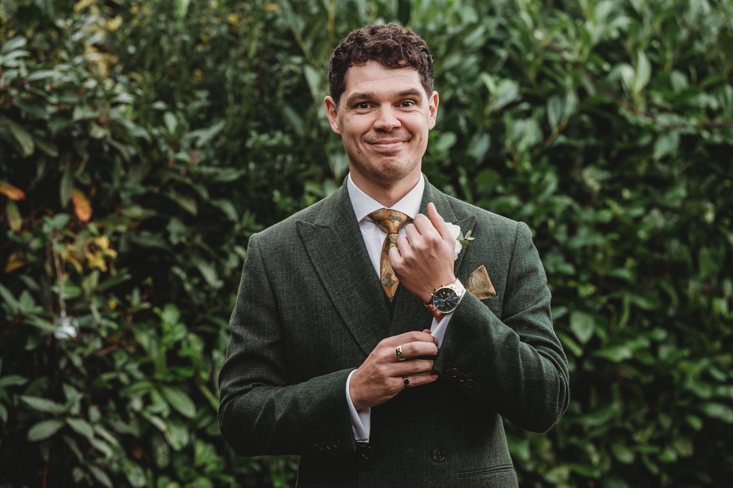 A groom in dark green suit adjusts his tie while smiling in an outdoor garden setting.